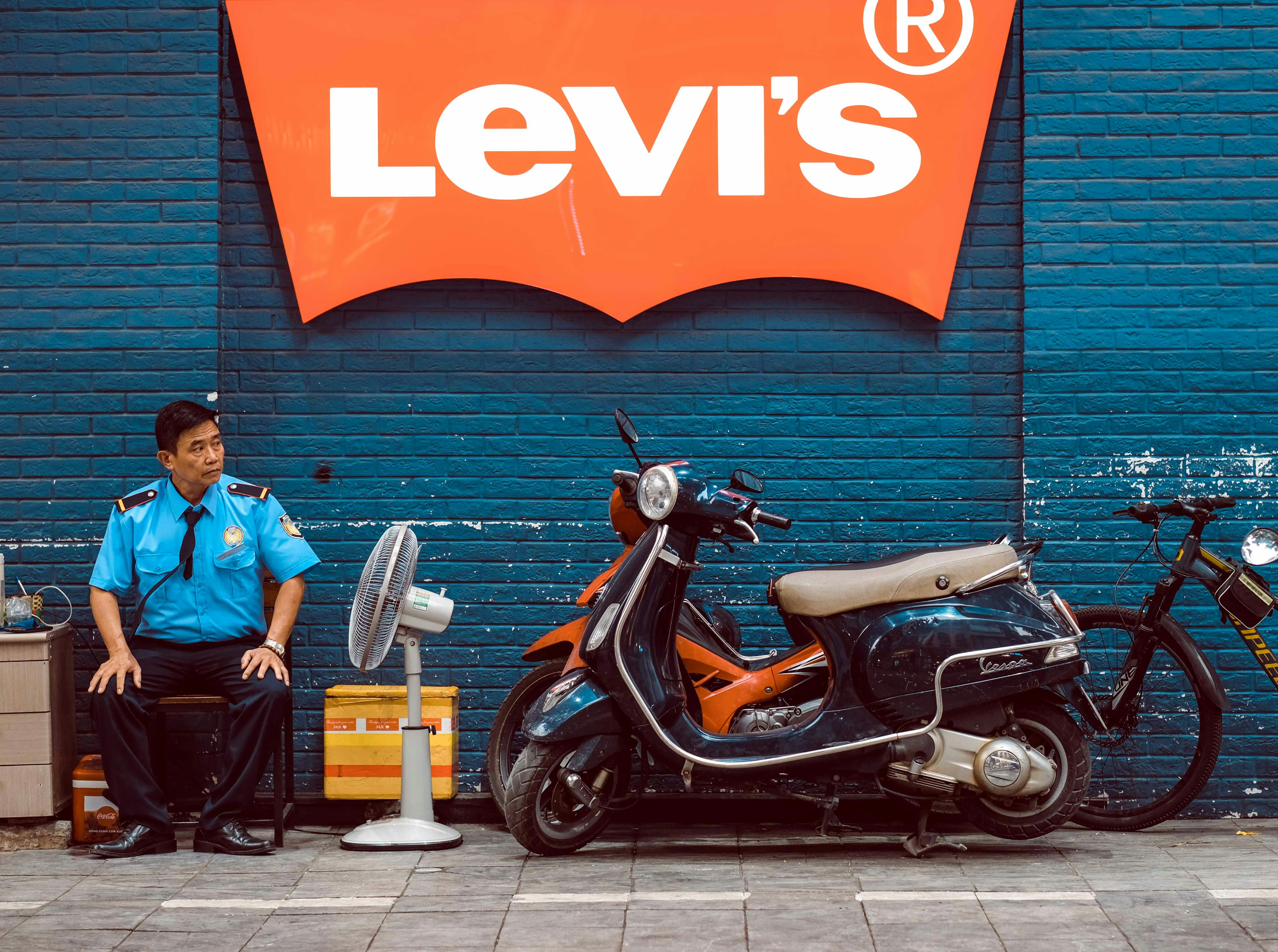 Man in uniform sits by fan next to scooter.