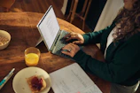 Woman typing on laptop at wooden table with breakfast.