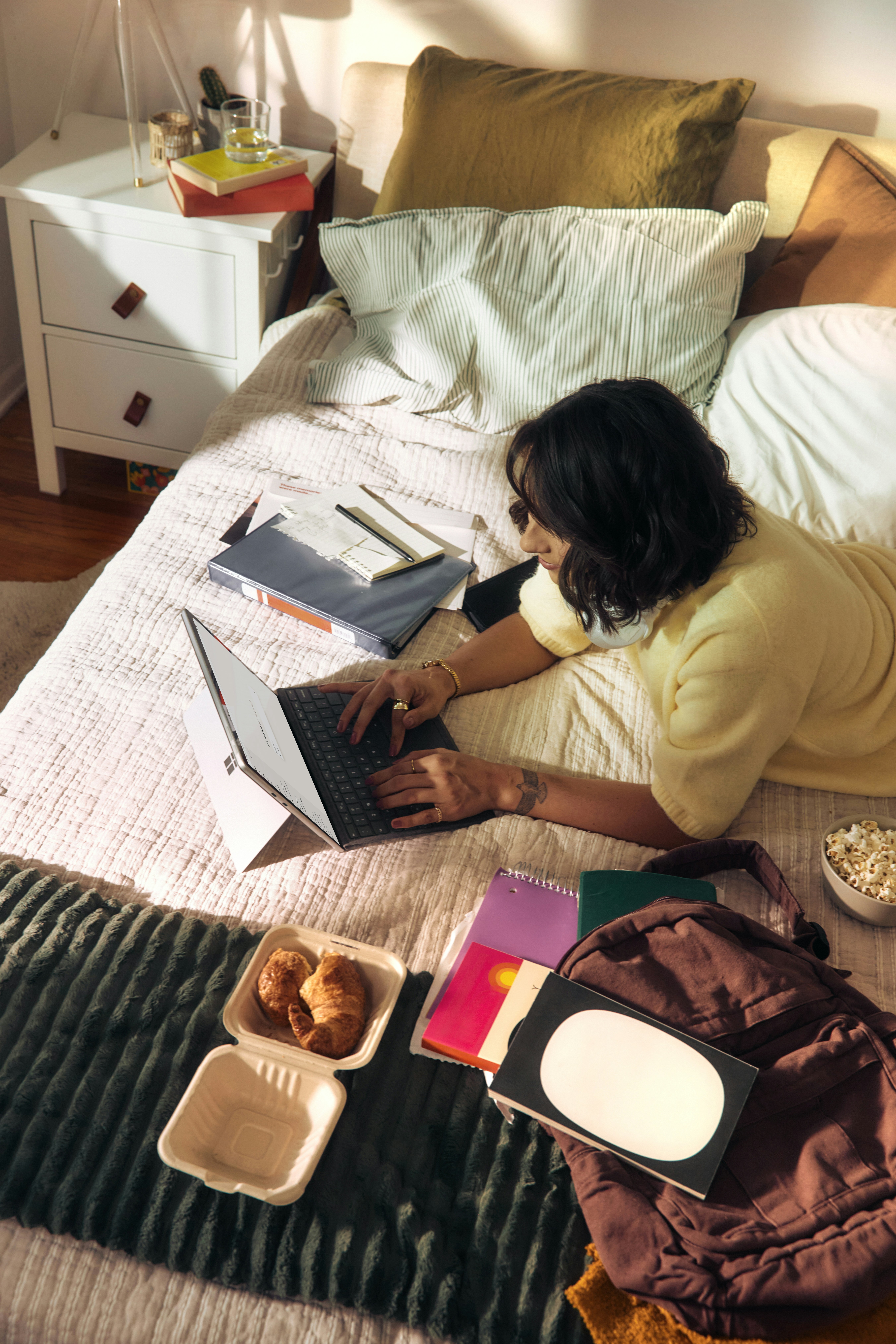 Woman working on laptop on bed with snacks and books.