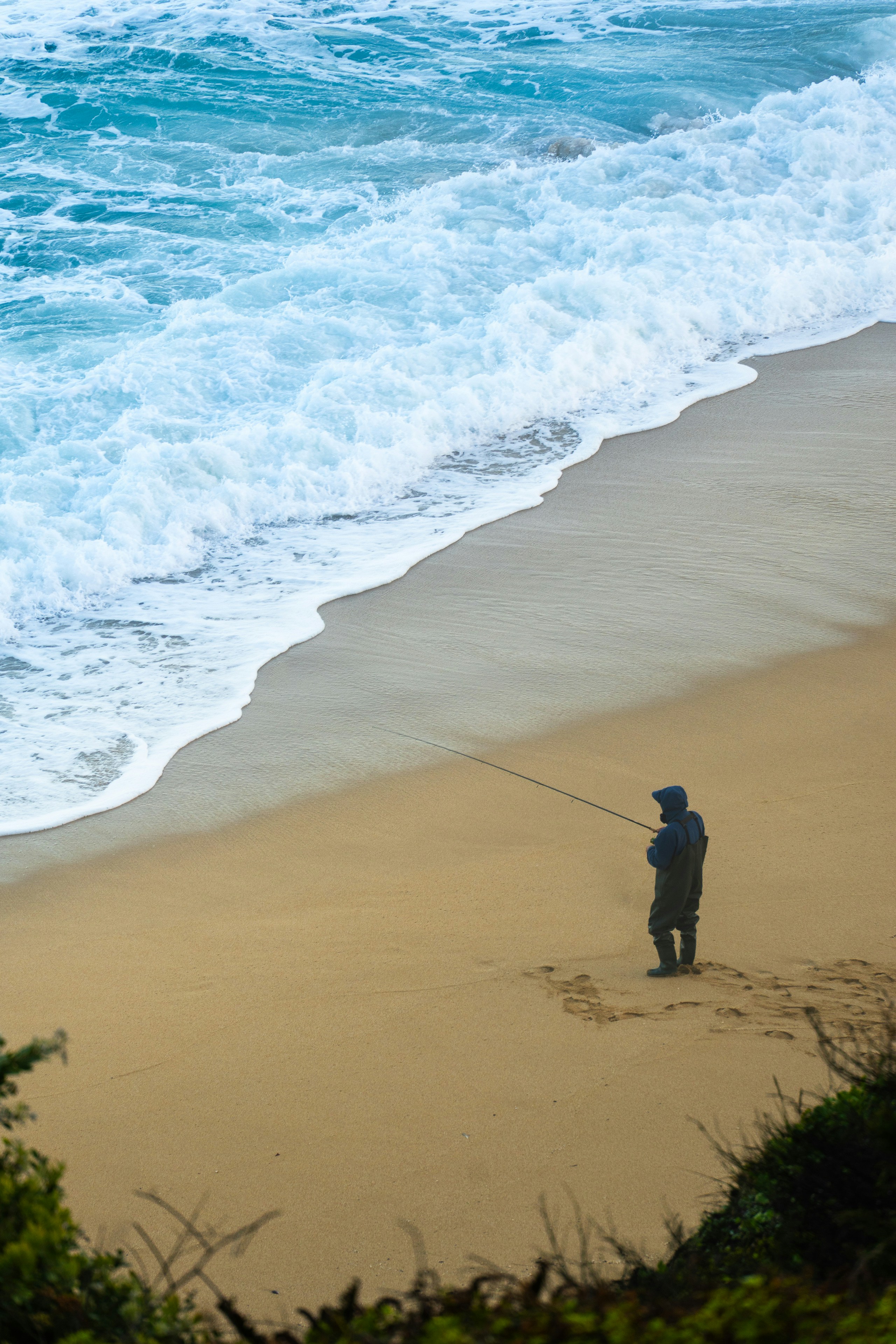 A lone fisherman casts a line on a sandy beach.