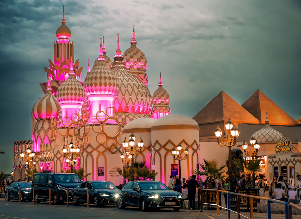 A pavilion with ornate onion domes lit pink at dusk at Global Village, with pyramid-shaped buildings beside it, lamp posts and parked cars in front under a dark cloudy sky