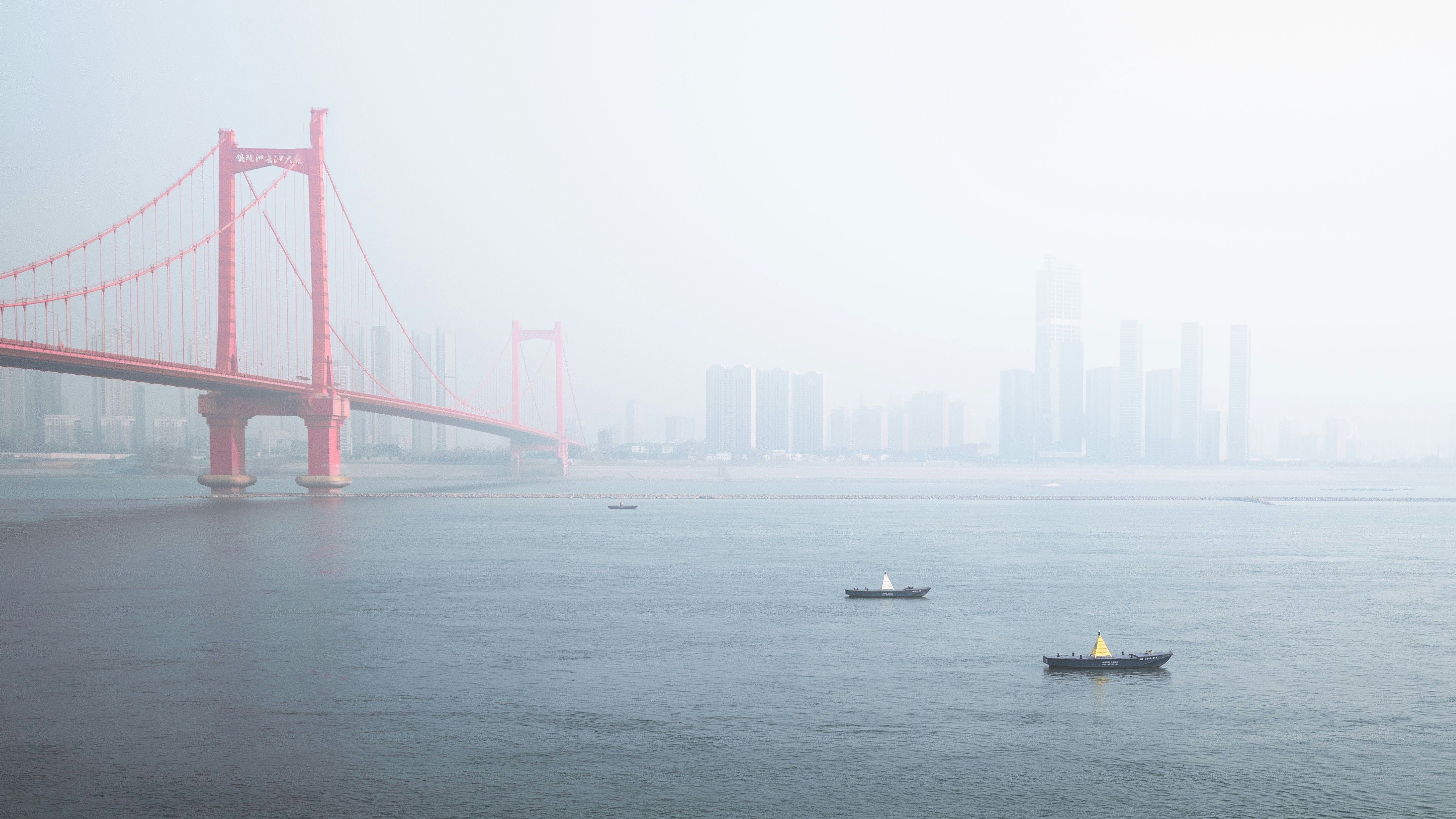 Red suspension bridge over water with boats and city skyline.