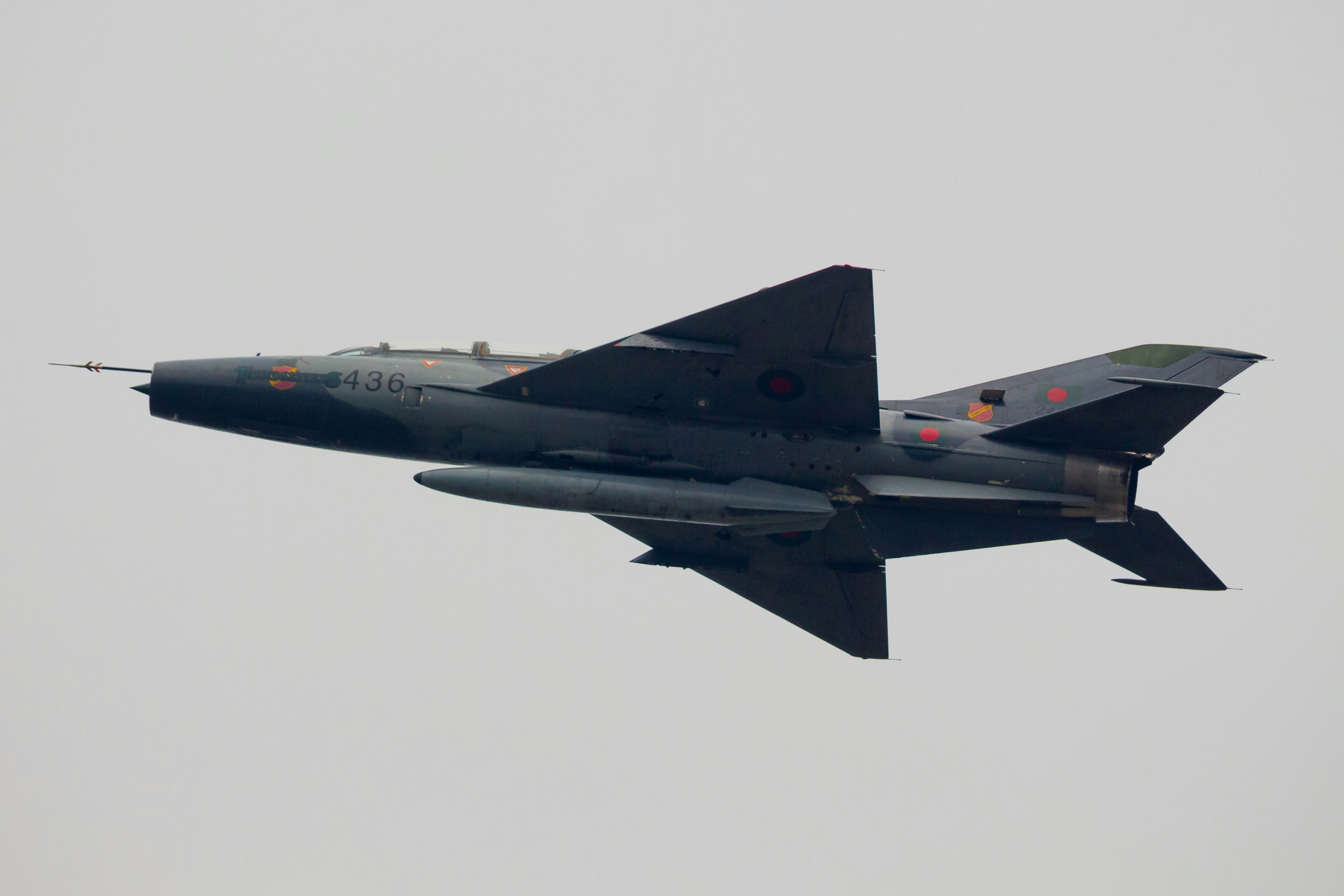 A dark gray fighter jet flies against a pale sky.