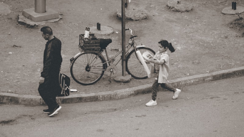 A child rides a bicycle on a paved neighborhood path next to an adult