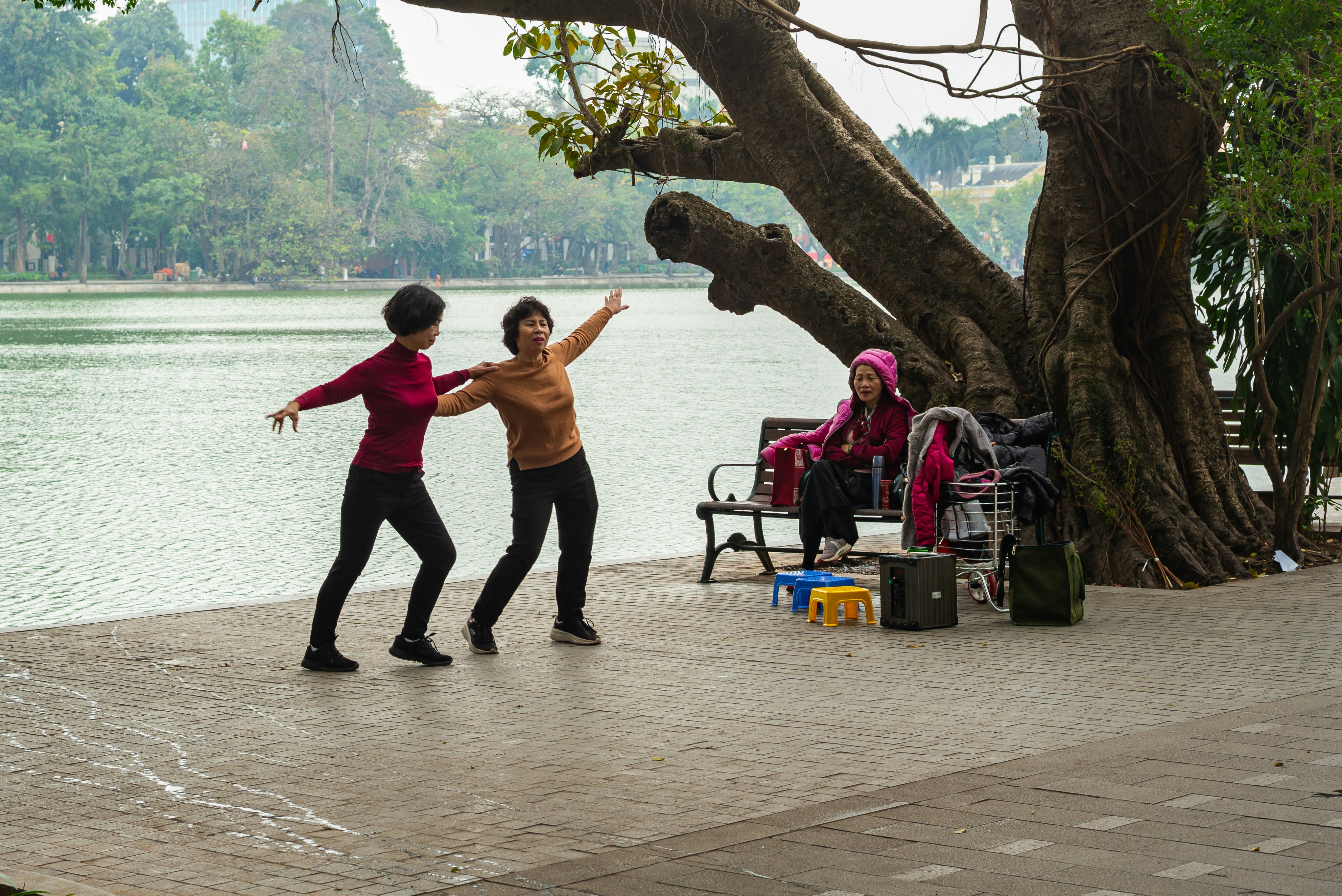 Two women dance by a lake, another sits nearby.