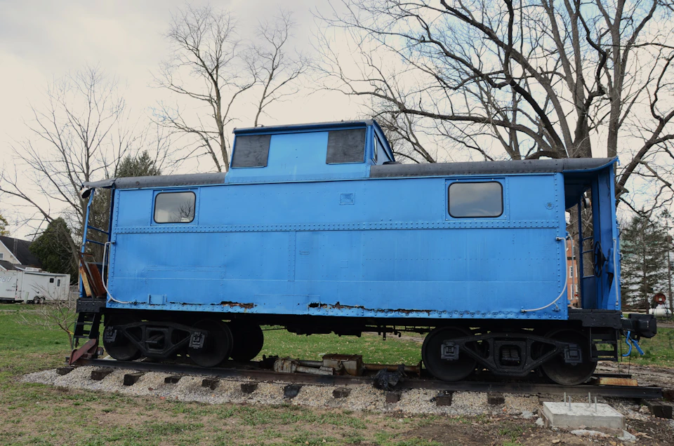 The image shows a blue train car with a black roof, parked on railroad tracks surrounded by grass and trees. The train car is positioned facing towards the right side of the frame, giving it an imposing presence against the backdrop of nature.