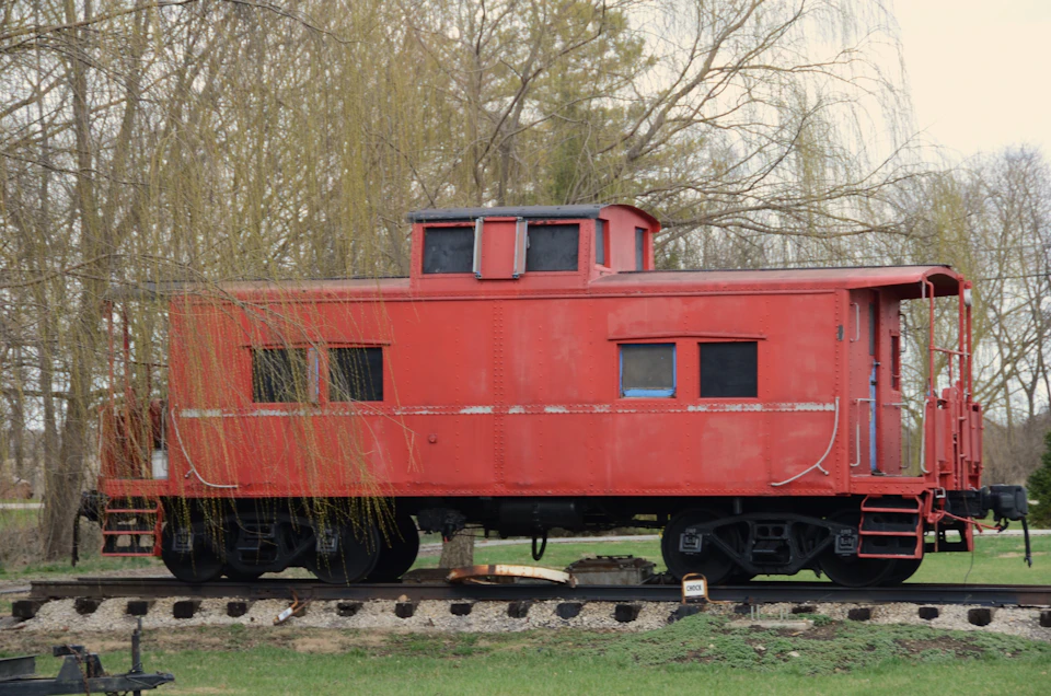 The image shows a red train car, specifically an old-fashioned steam locomotive, parked on railroad tracks surrounded by trees and grass. The train car is positioned facing towards the right side of the frame, with its caboose visible at the front. The scene conveys a sense of nostalgia and history as the train car rests in a serene outdoor setting.