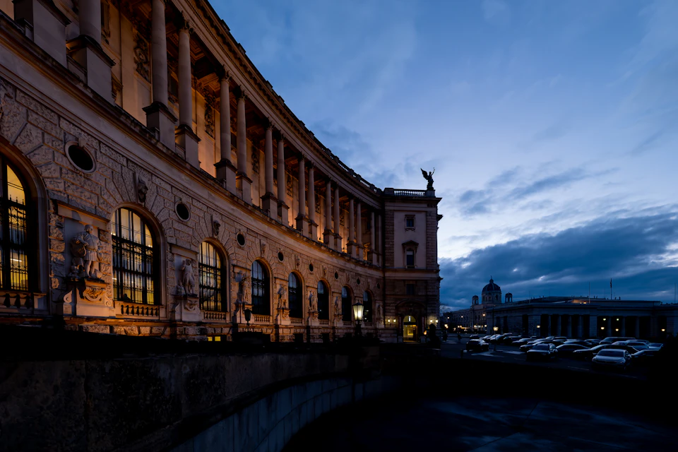 The image depicts a large, ornate building with a curved facade and numerous columns, situated on a street corner. The building is bathed in warm light from its windows, creating an inviting atmosphere. A statue stands atop the building, adding to its architectural charm. In front of the building, there are several cars parked along the street, suggesting that it may be a popular location or a busy area. The sky above is filled with clouds, indicating that the photo was taken during the evening hours.