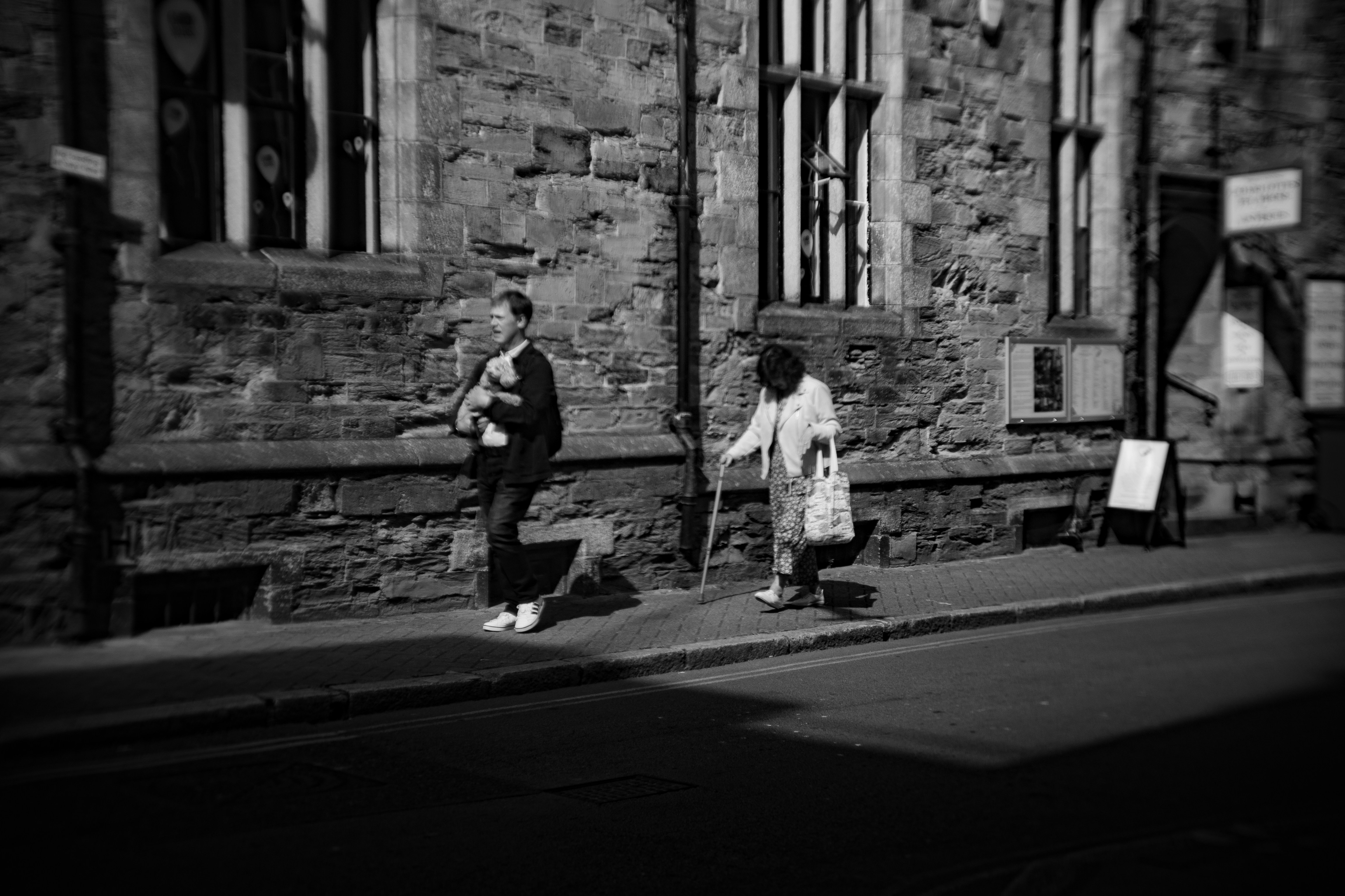 Two people walk on a sidewalk beside a stone building.