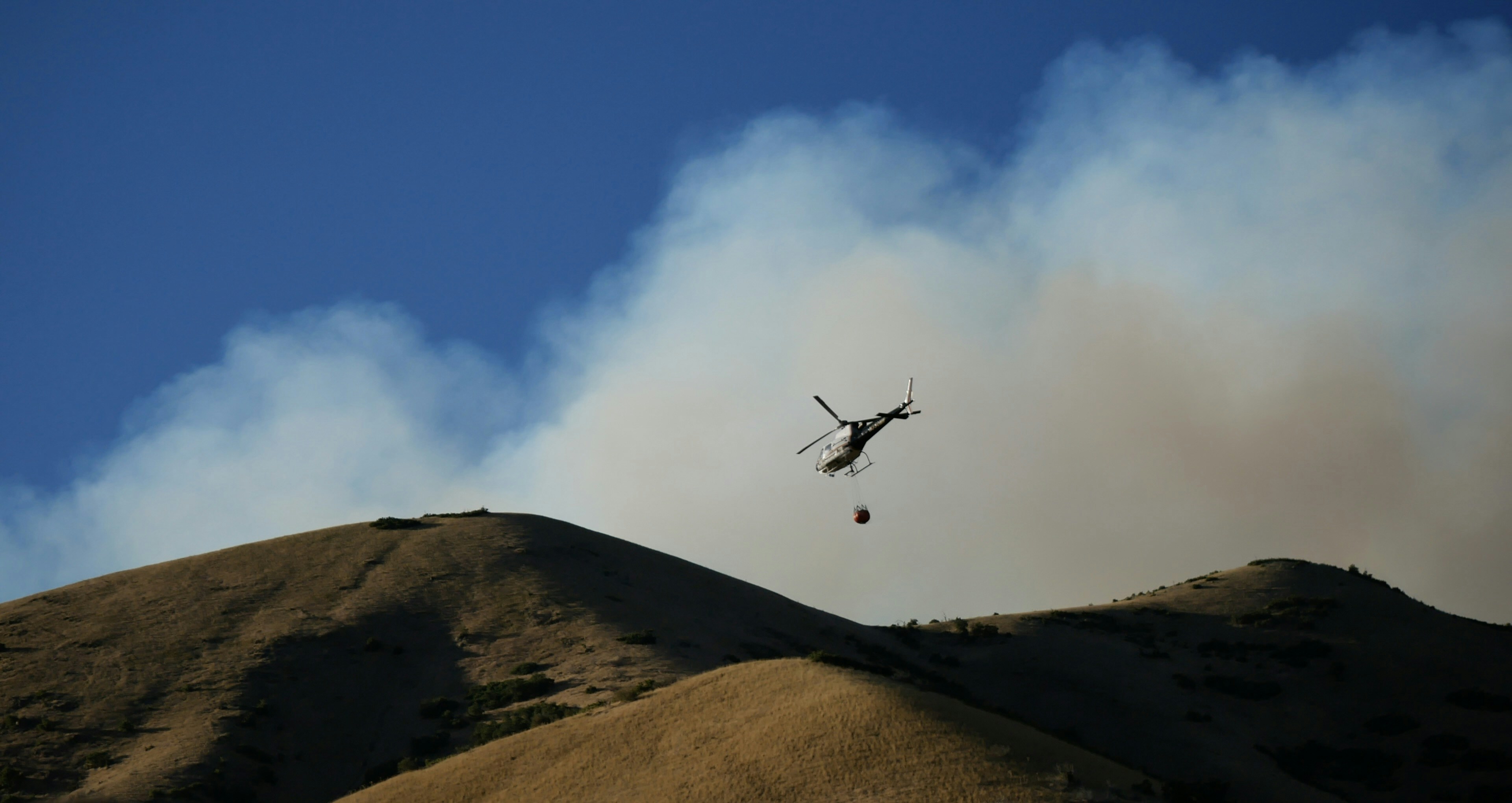 Ein Hubschrauber wirft Wasser auf ein Feuer am Hügel.