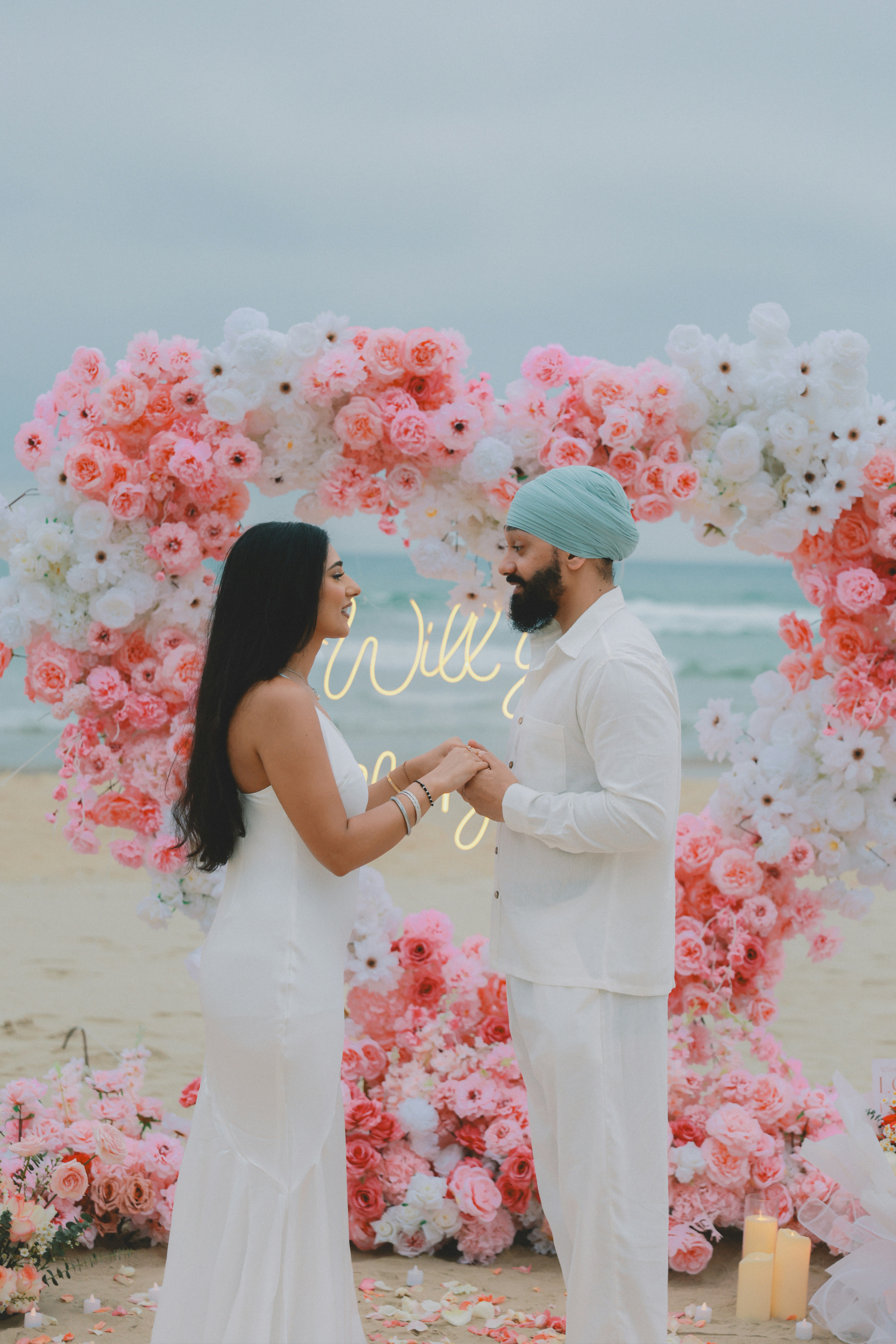 Un couple se tenant la main près d’une arche en forme de cœur floral sur la plage