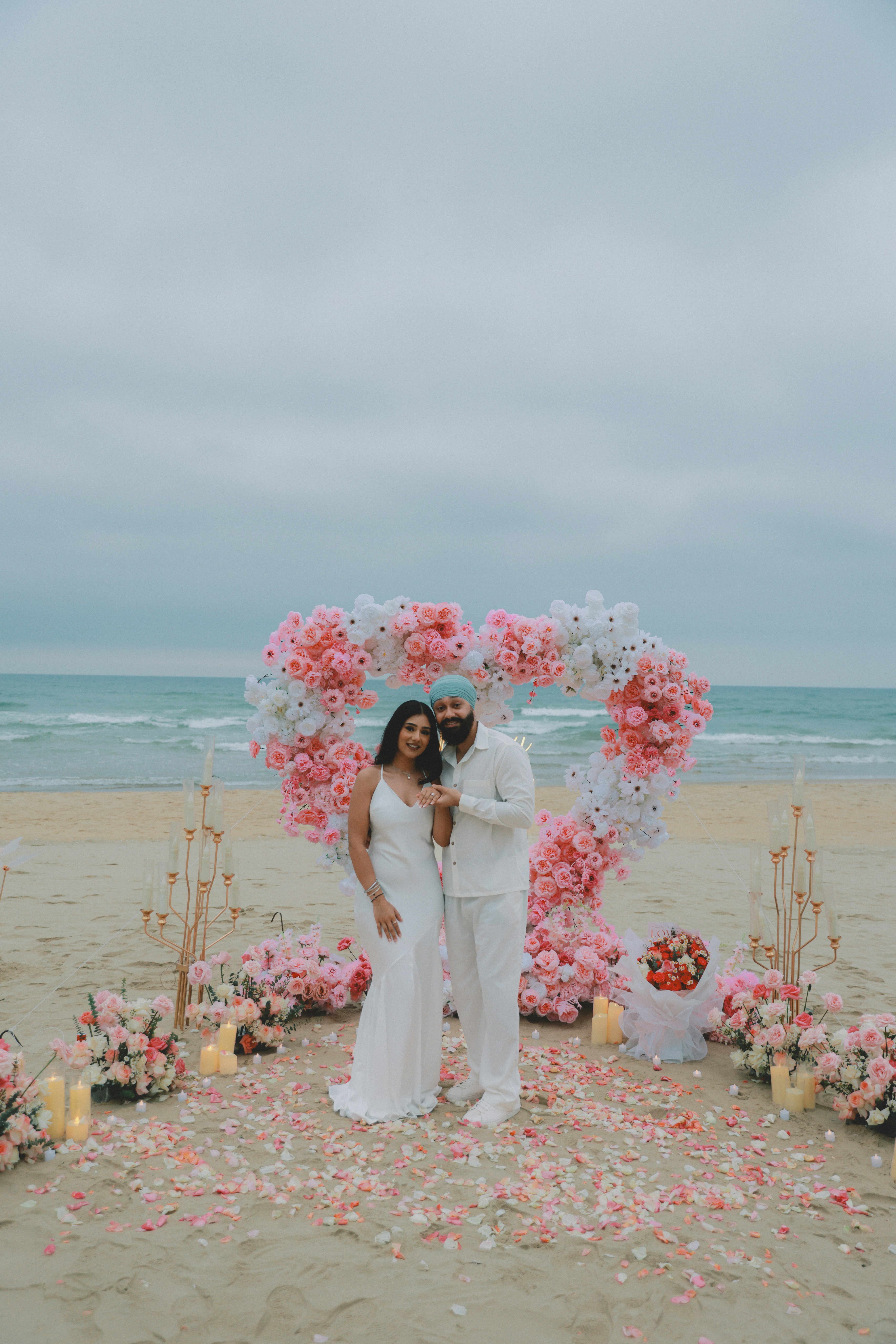 Un couple pose près d’une arche florale en forme de cœur sur une plage.