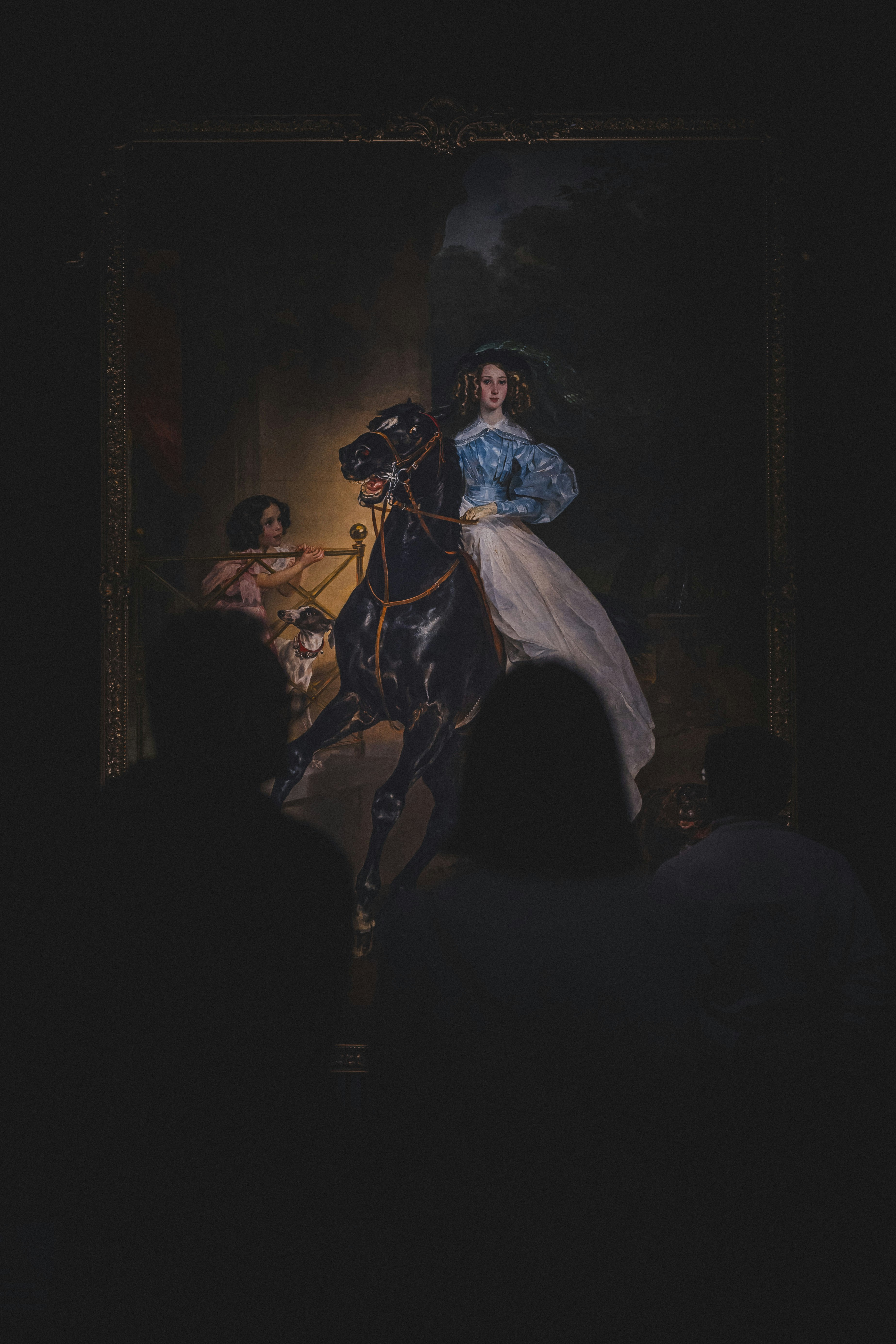 This captivating photograph features a majestic painting by Karl Bryullov displayed in the New Tretyakov Gallery in Moscow. The artwork, illuminated against a dark background, portrays a graceful woman elegantly dressed and seated upon a powerful black horse, accompanied by a young girl and a dog. Silhouetted visitors in the foreground add a sense of awe and engagement, drawing viewers into the dramatic, historical scene.