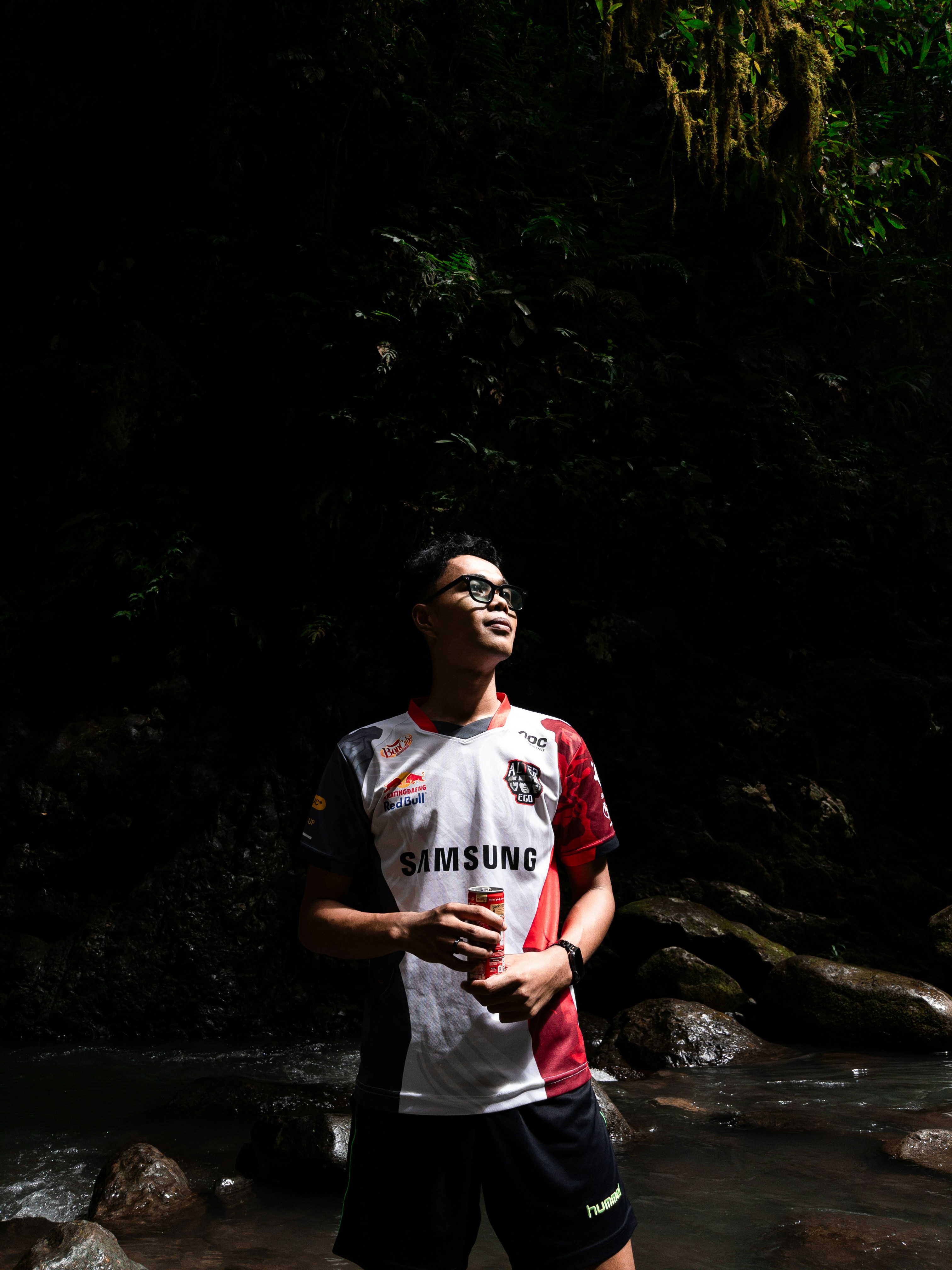 Pria berbaju berdiri di tepi sungai di hutan.