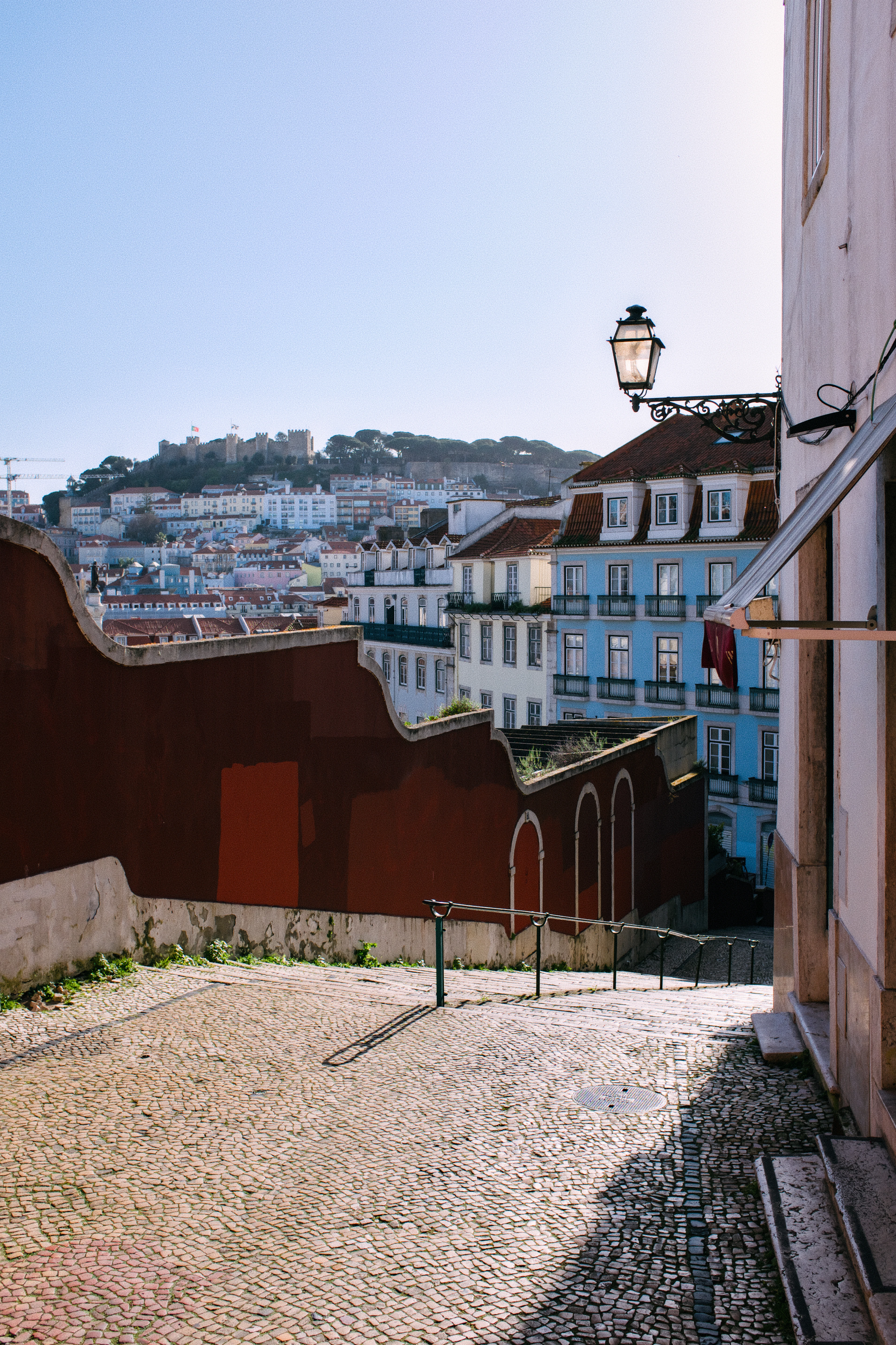 A quiet cobbled terrace on the slopes of Alfama in Lisbon in low sun, a black iron street lantern overhead and a deep red wall in the foreground, looking across pale and blue-tiled townhouses to the ramparts of Sao Jorge Castle on its wooded hill