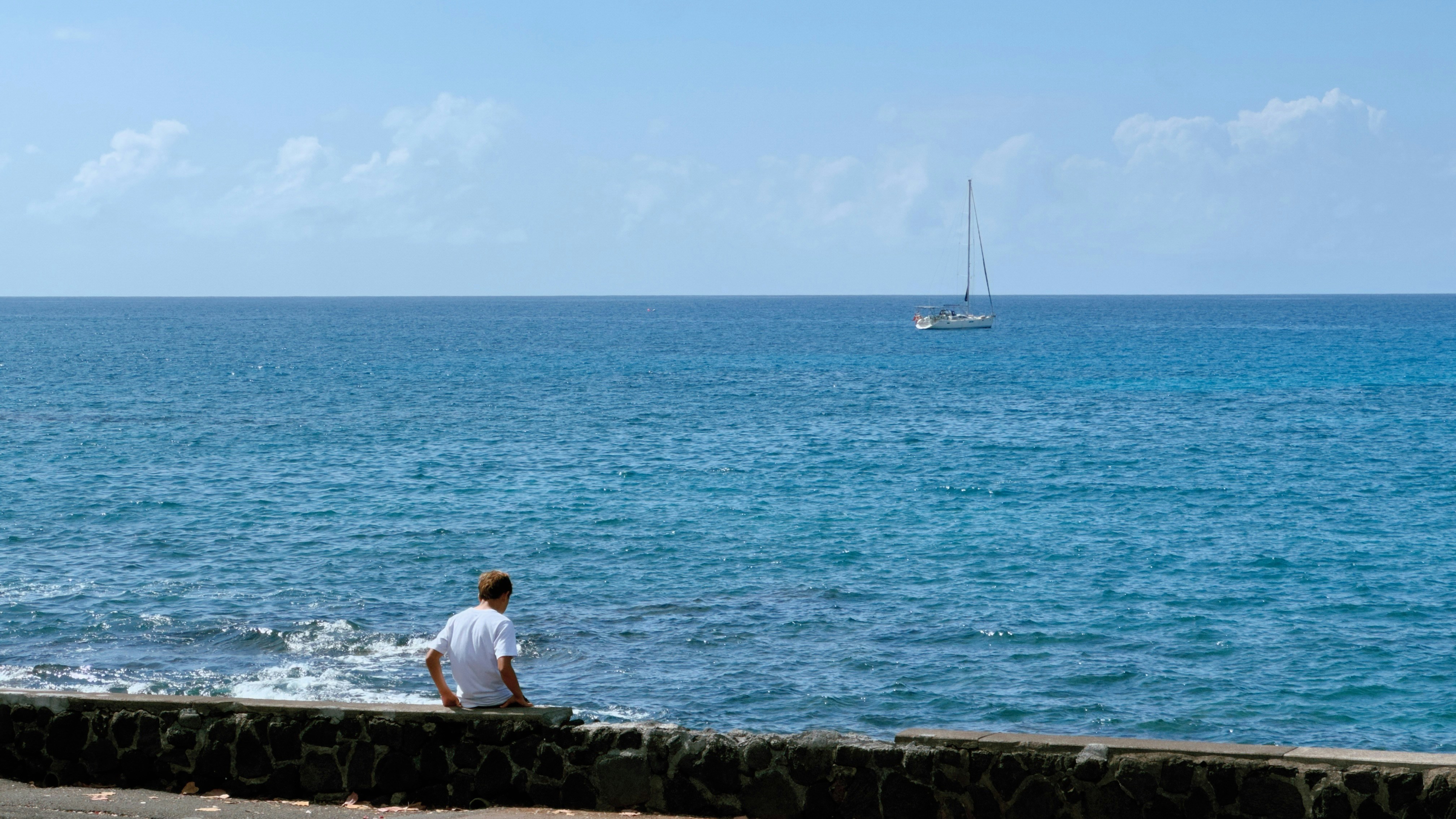 Hombre sentado en la pared con vistas al océano con velero