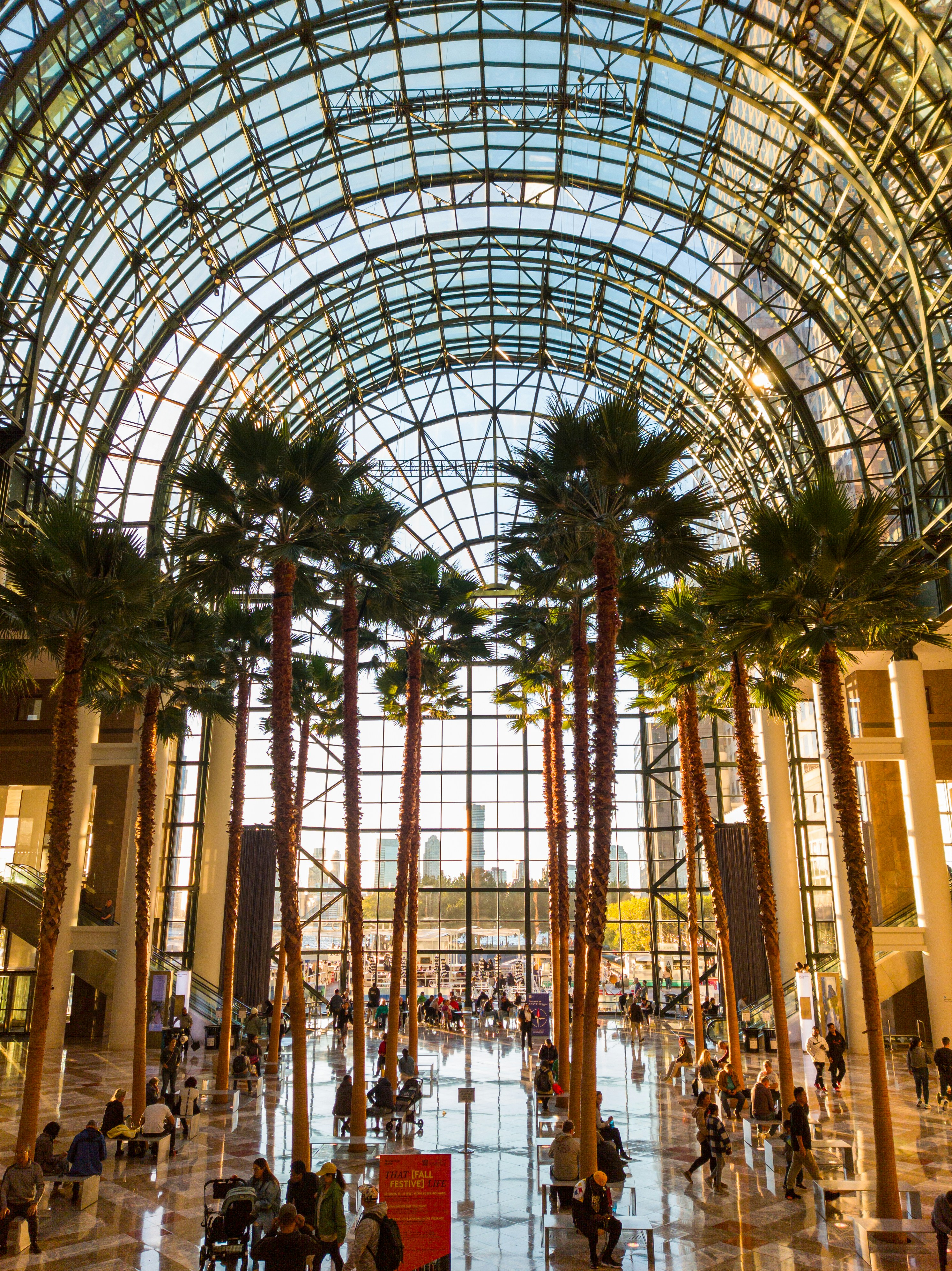 웅장한 유리 아트리움 안에 키 큰 야자수들이 있고, 사람들이 모여 있다