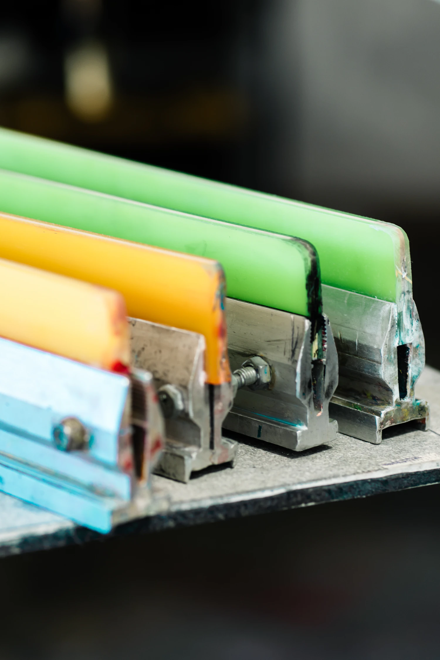Squeegees with green, yellow and blue blades lined up on a rack in the print studio