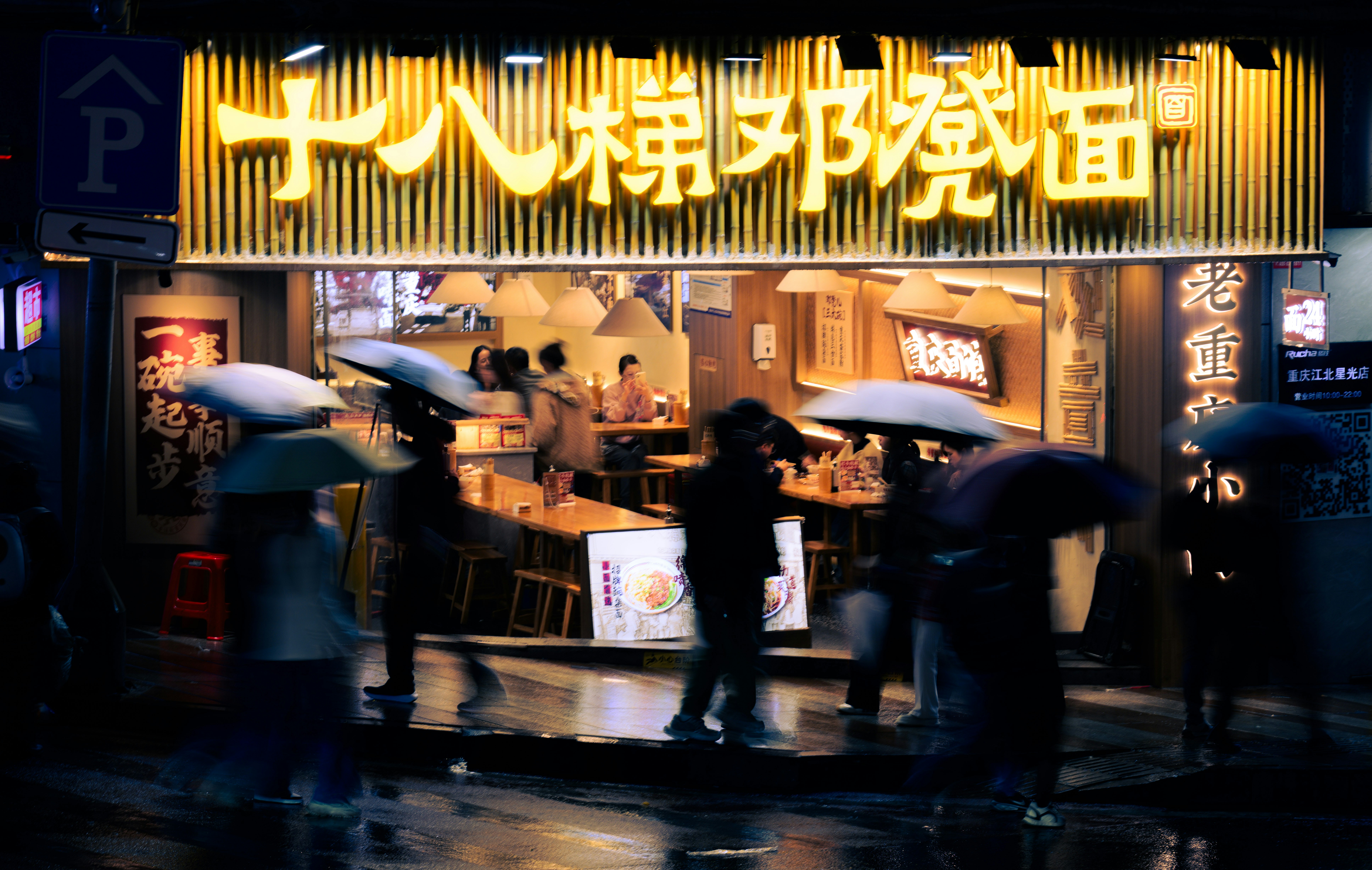 People with umbrellas walk past a noodle shop at night.