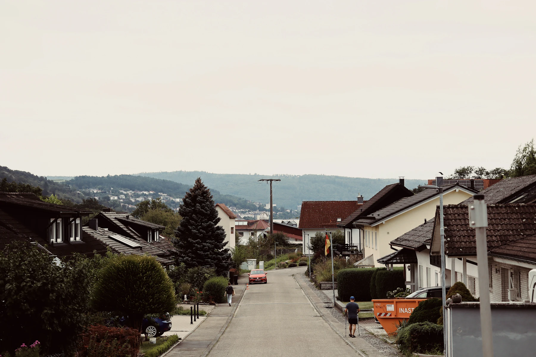 A quiet suburban street running downhill between houses and hedges, with green hills beyond