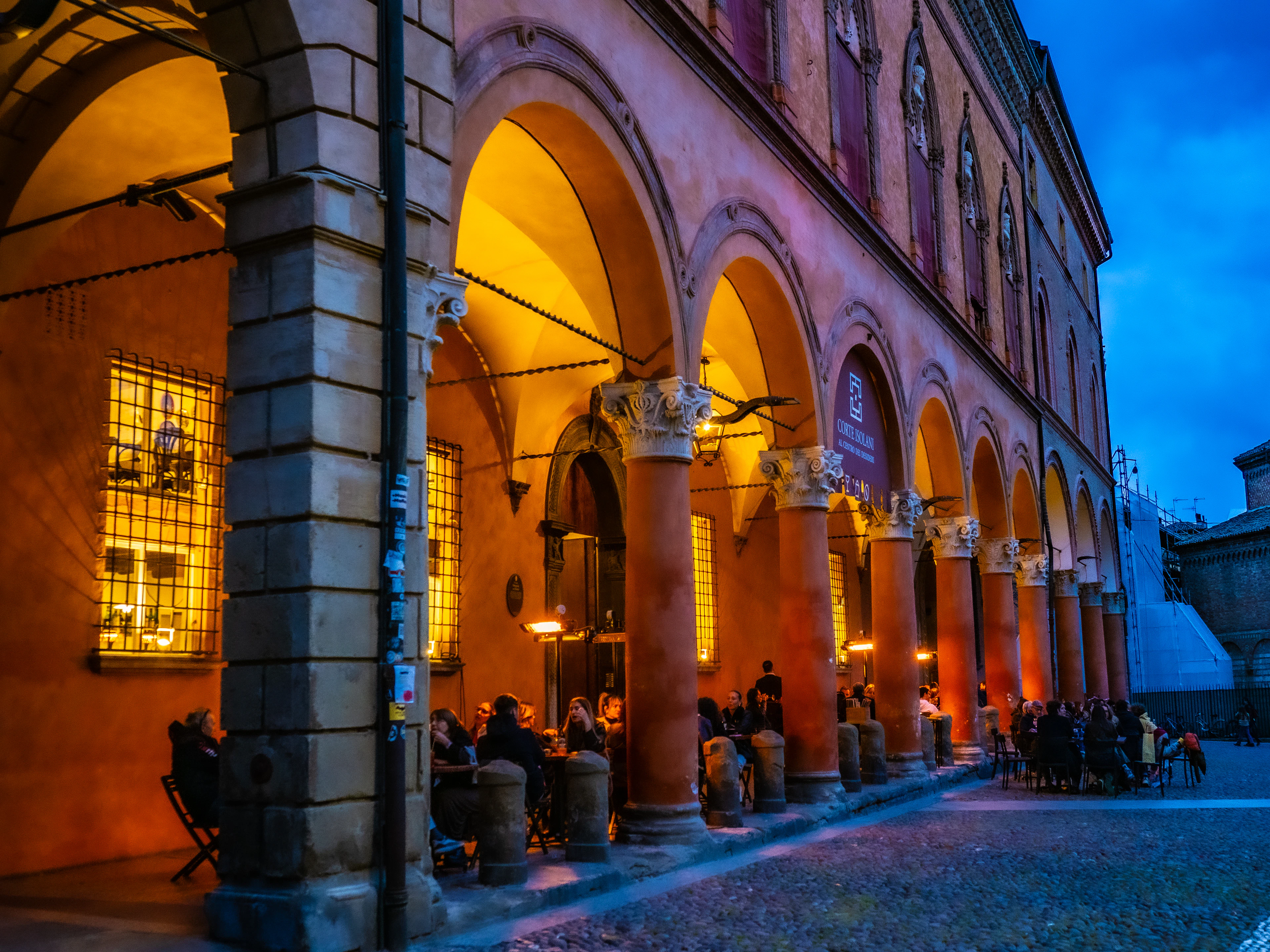 People eating and drinking at tables under the lit arches of a Bologna portico at dusk