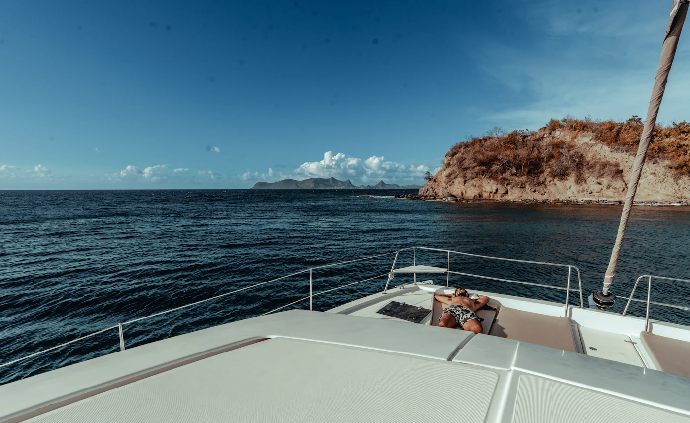 The wide foredeck of a sailing boat lying at anchor over deep blue water, with a rocky coast beyond.