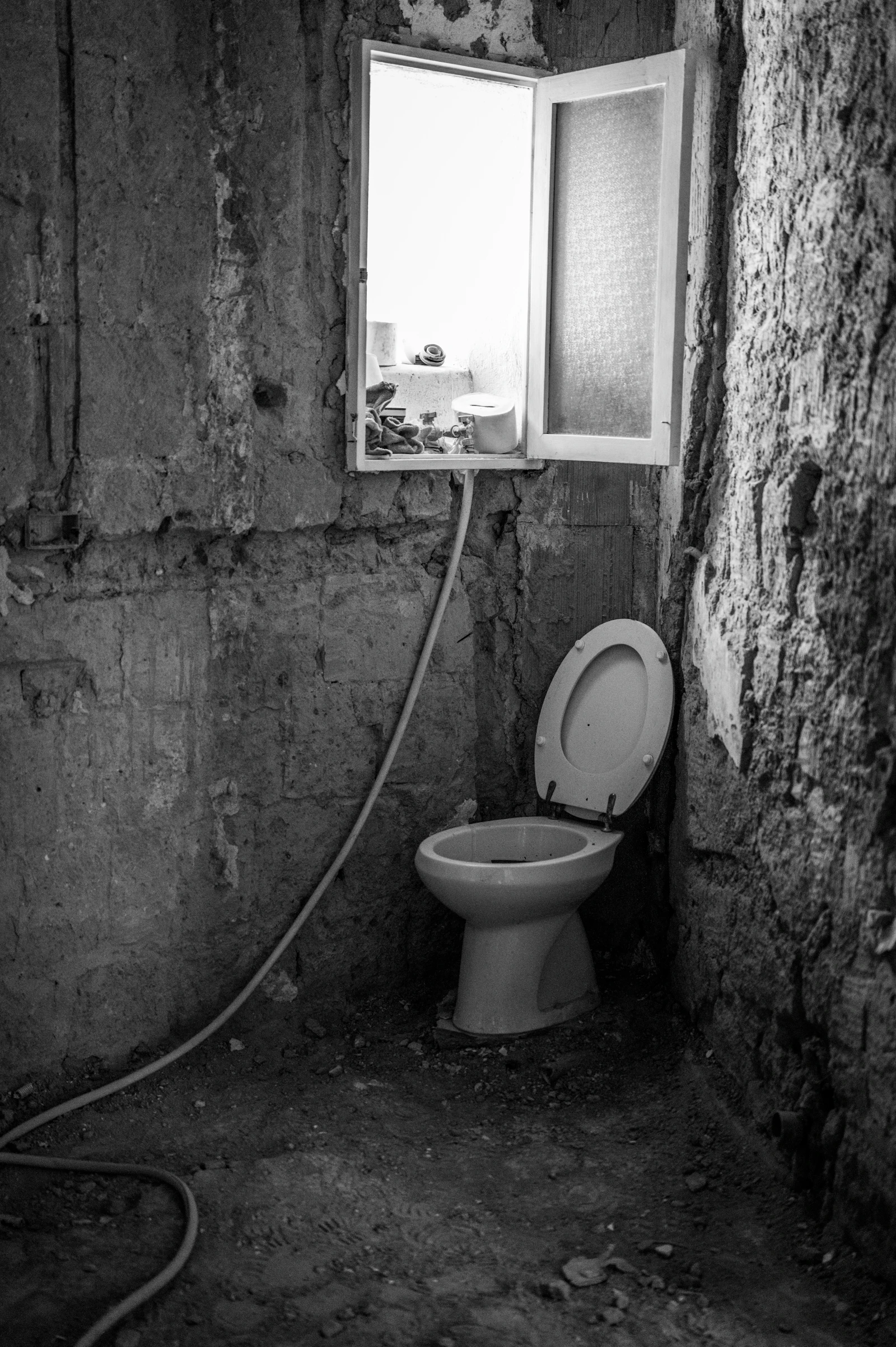 Unfinished toilet room with rough masonry walls, exposed plumbing, an open window and a standalone toilet.