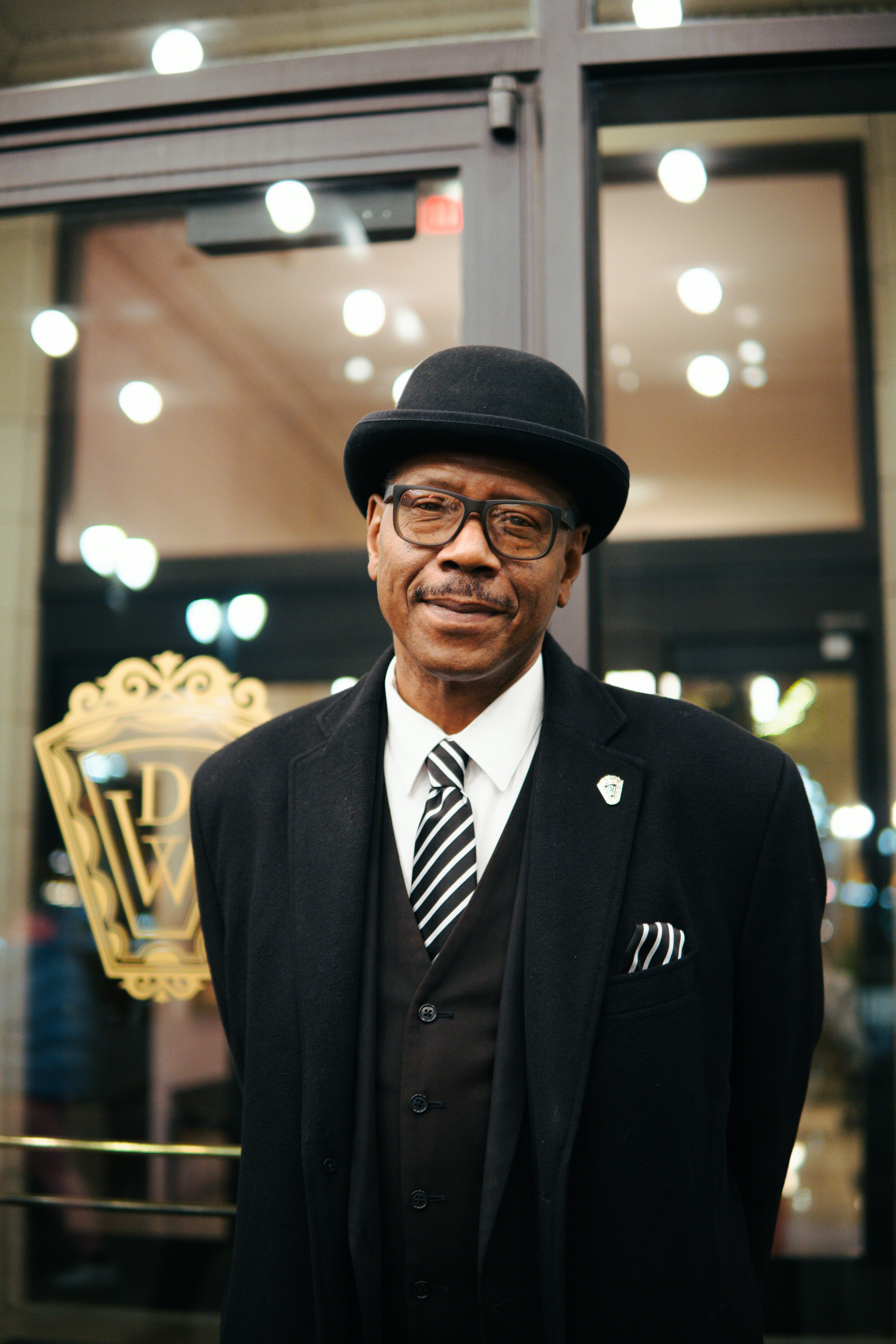 Man in a bowler hat and suit stands outside building.