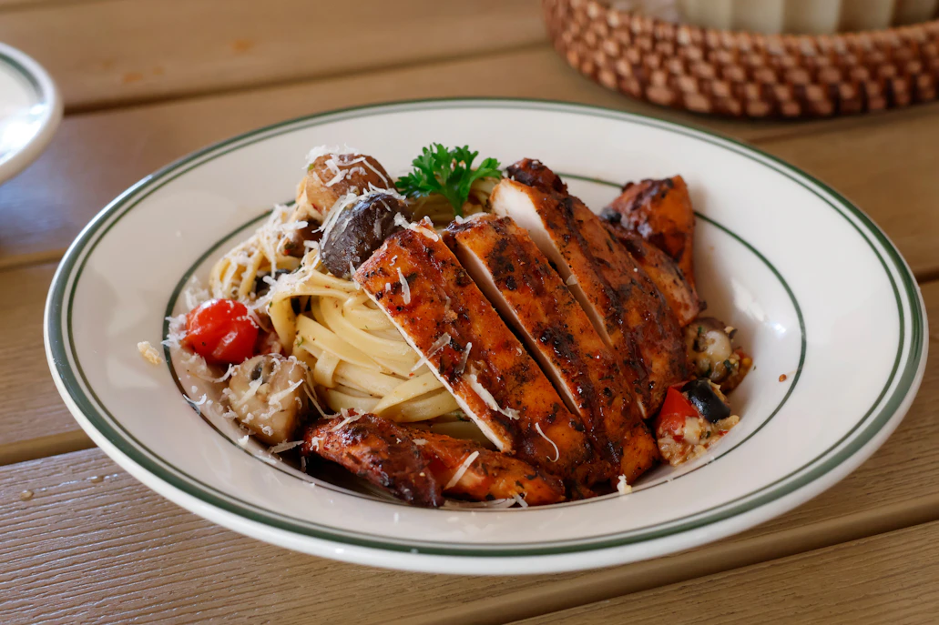Sliced grilled chicken breast over pasta with vegetables.
