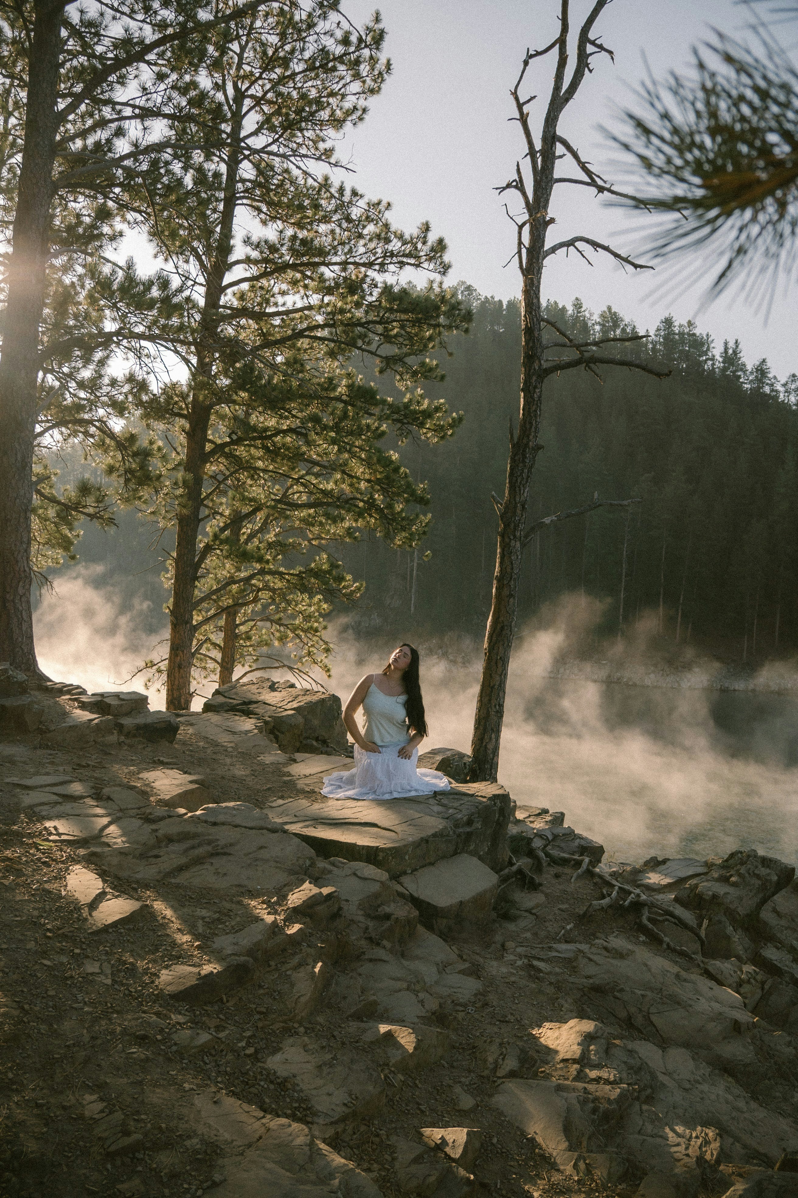 Woman in white dress sits on rocky shore with mist.
