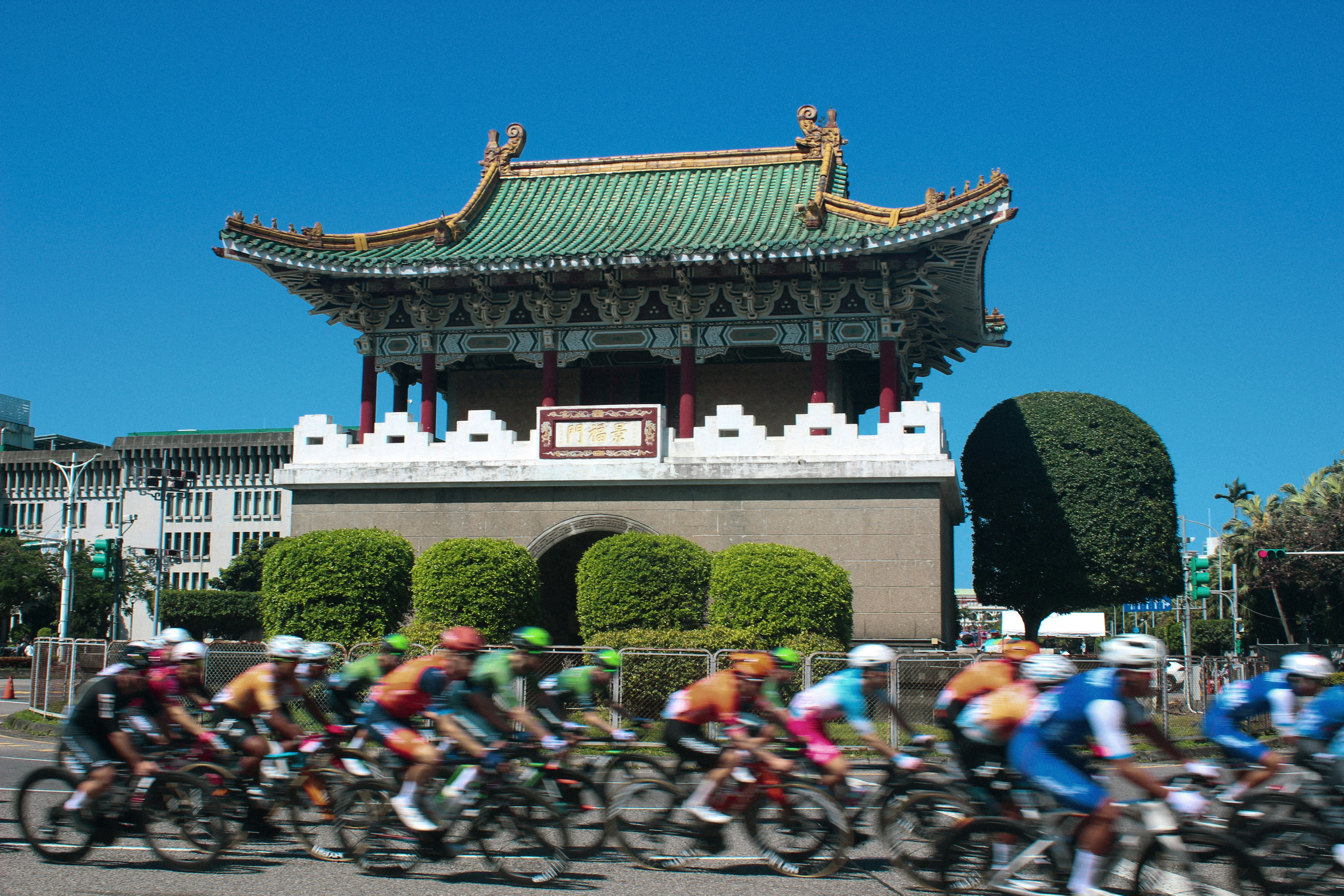 Cyclists race past a traditional building under a clear sky.