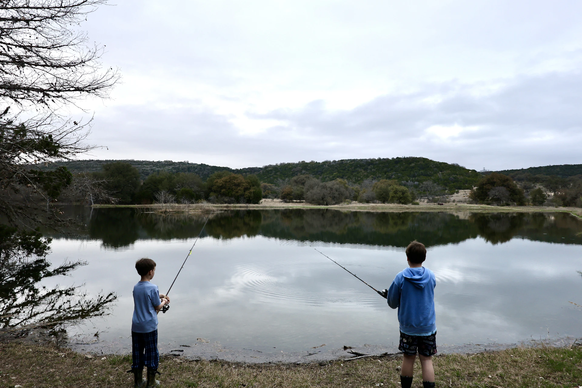 Two boys fishing by a calm lake with hills.