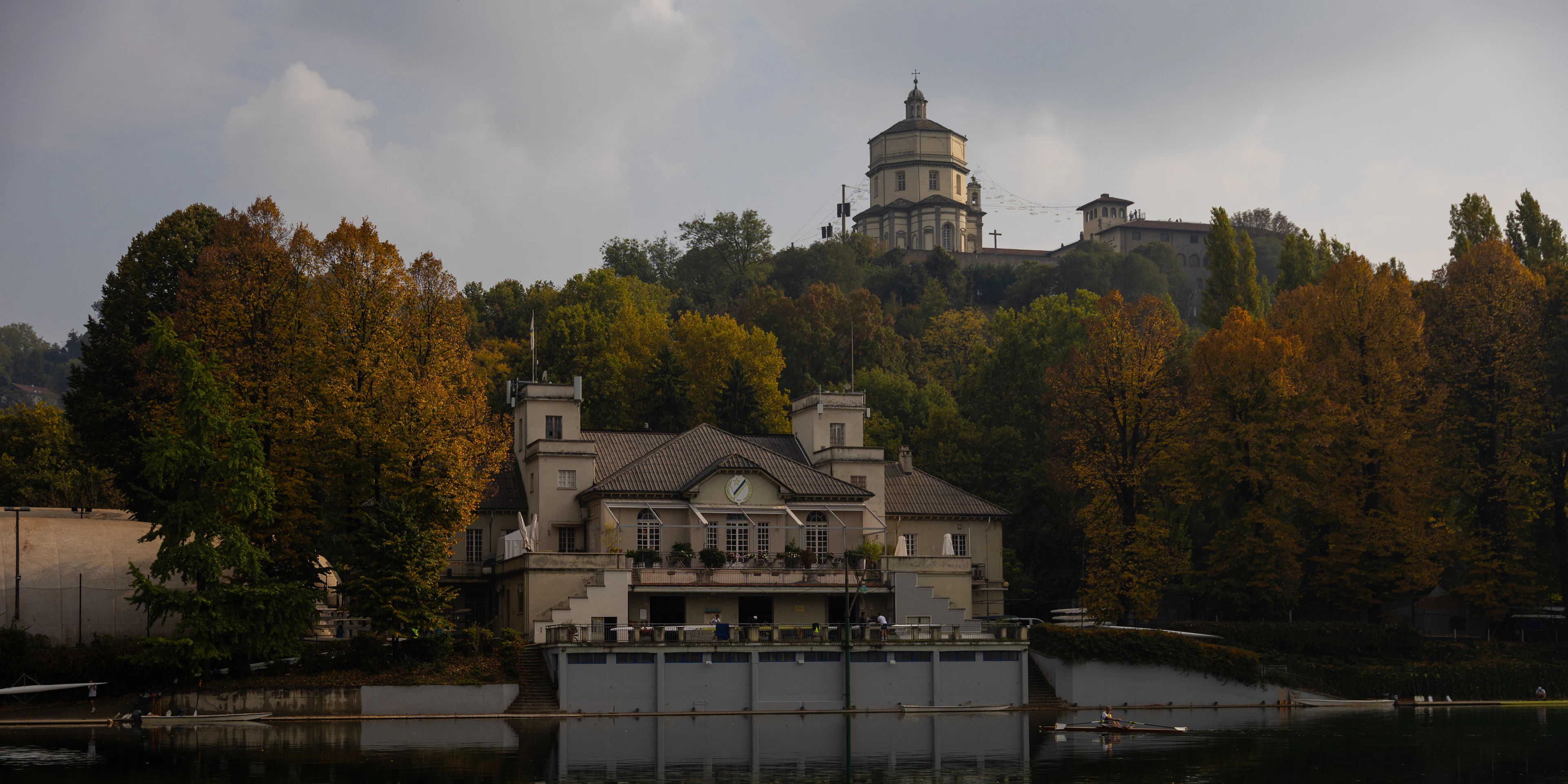 The domed church on the Monte dei Cappuccini hill above the River Po in Turin, seen from the far bank through gold and green autumn trees with a riverside rowing club on the water below.