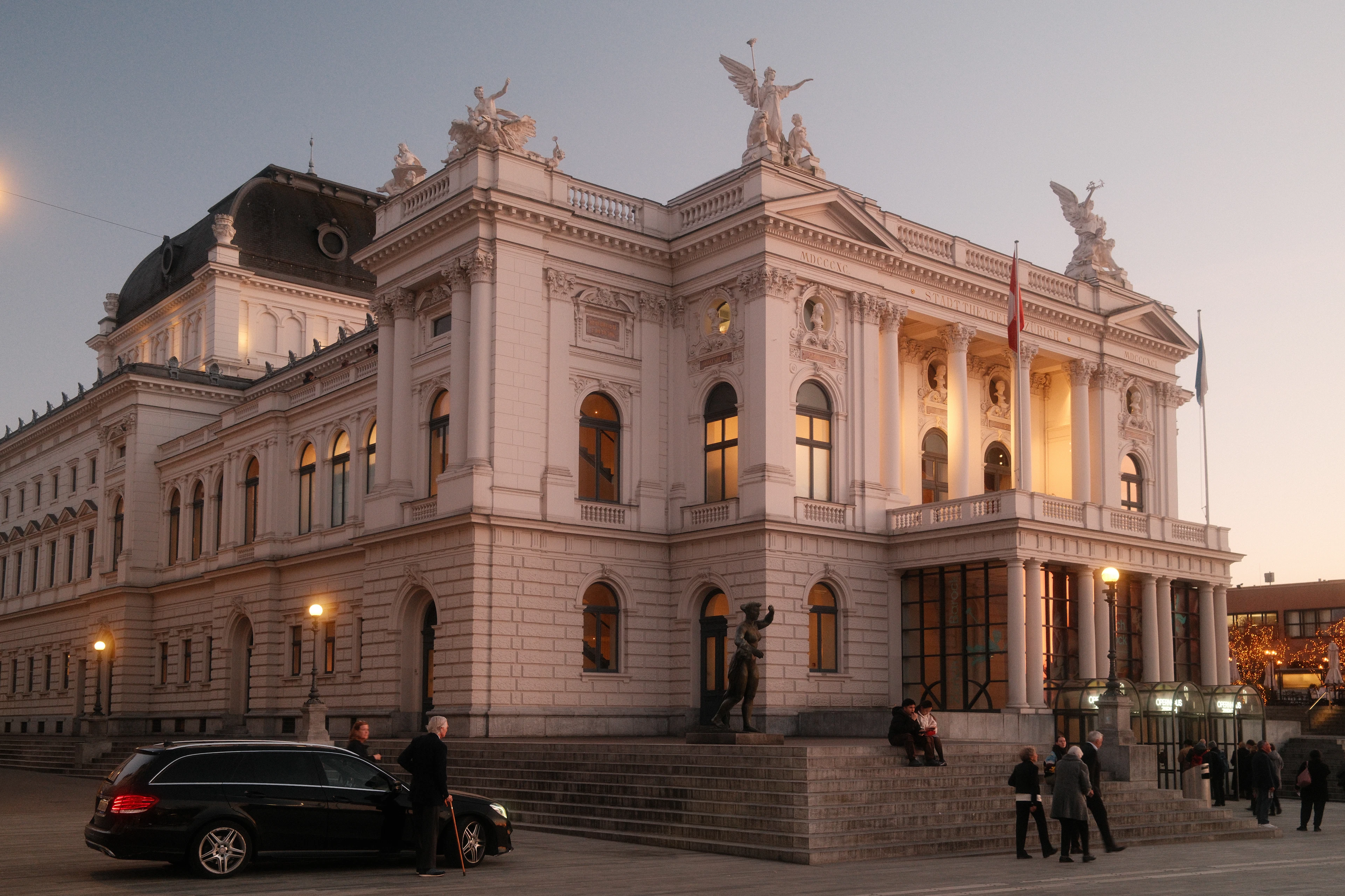 Zurich's neoclassical Opera House at dusk, its columned facade and winged rooftop statues pale against a violet sky while the foyer windows glow gold and a small crowd gathers on the steps before a performance