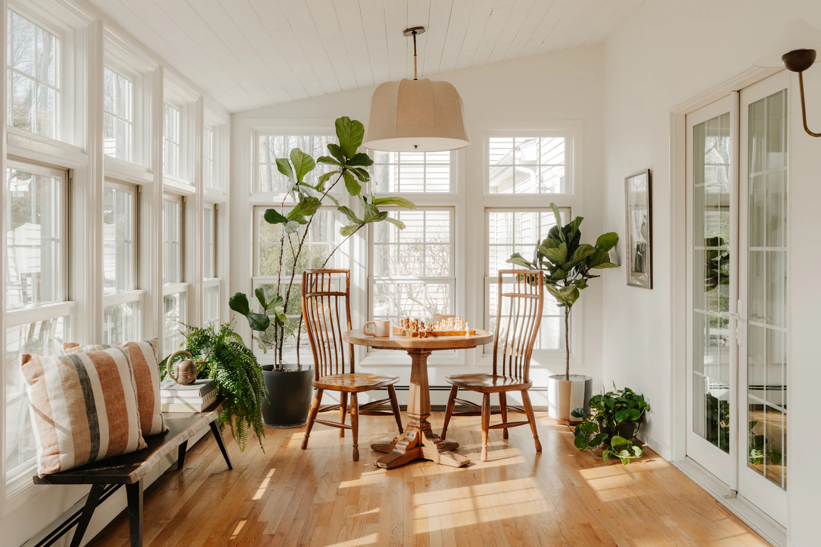 A sunroom in a house: windows on every side, morning light, a round wooden table under a pendant lamp, and plants