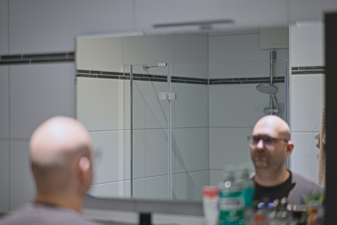 Bald man shaving his head with an electric shaver in front of a bathroom mirror