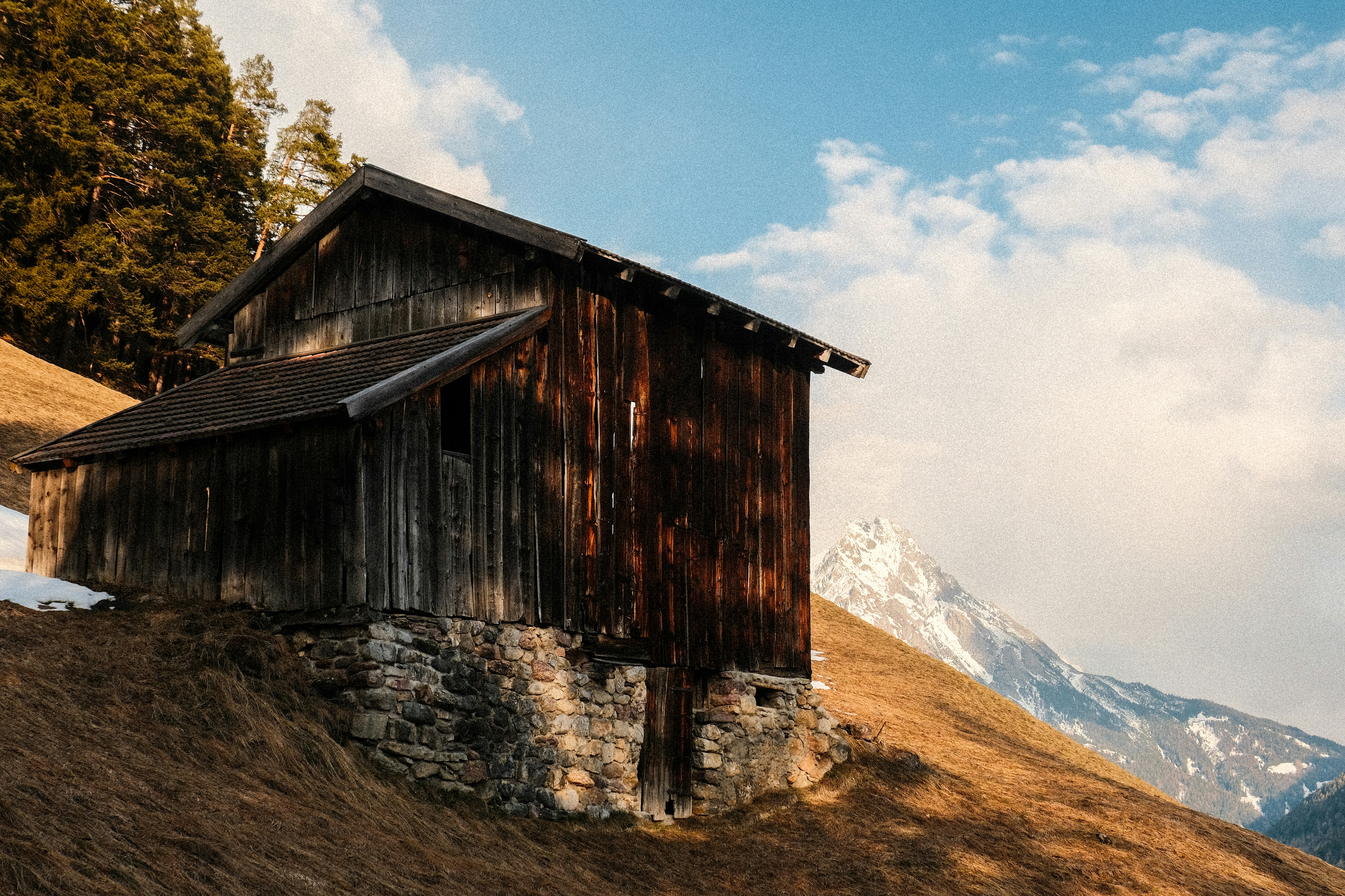 Old wooden barn on a grassy hillside with mountains.