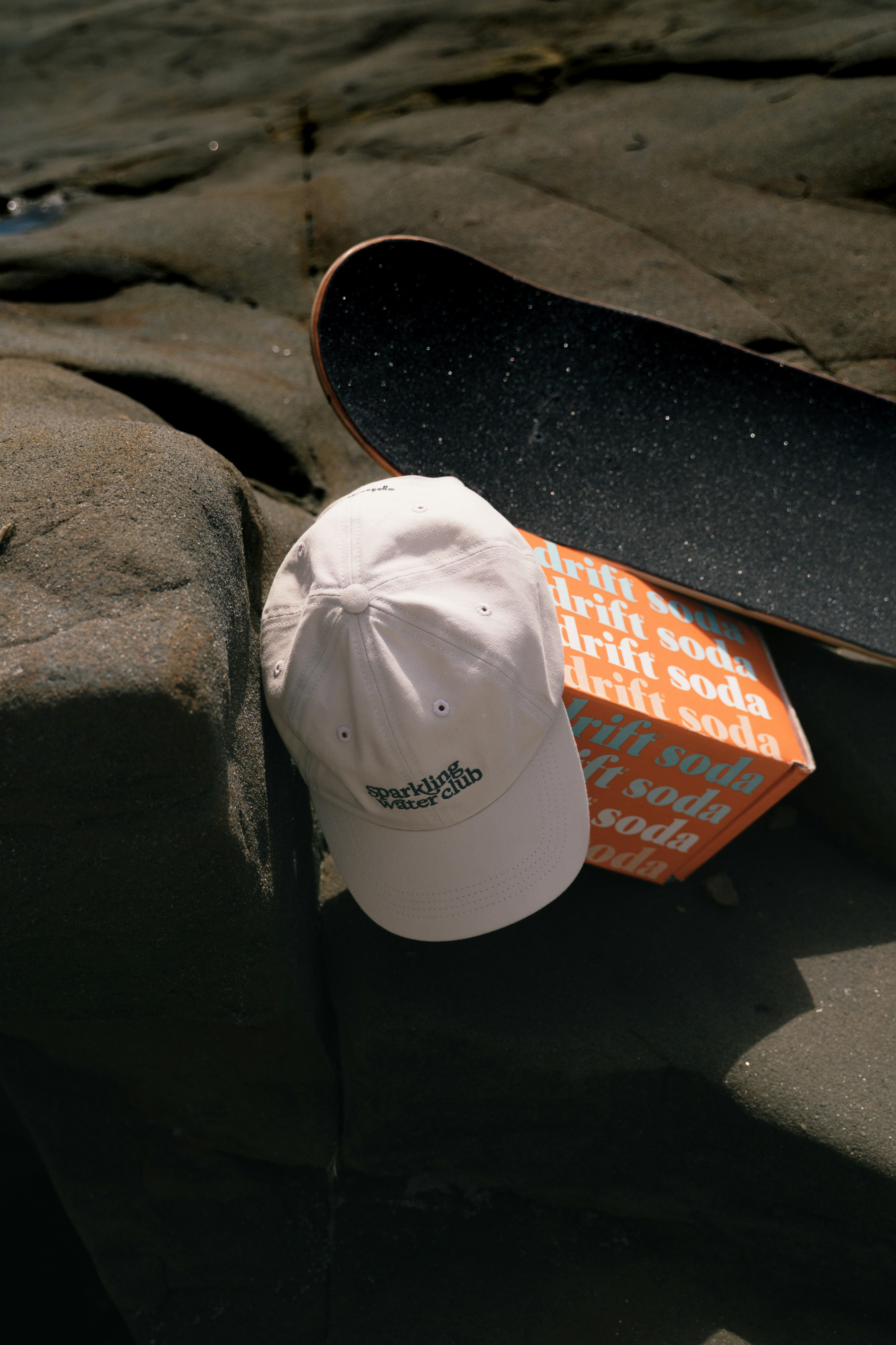 Un monopatín, una gorra y una caja de refrescos sobre piedras.
