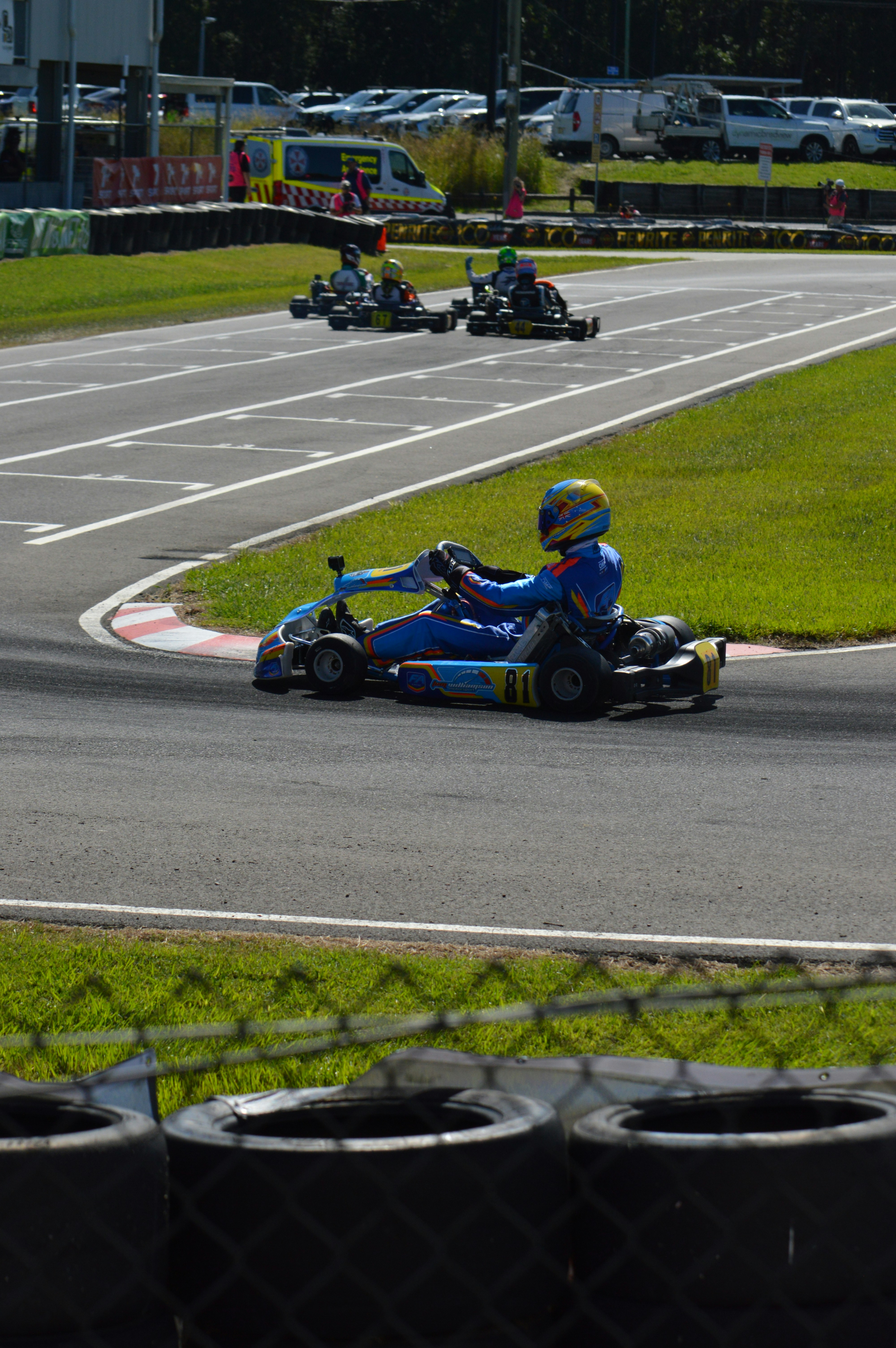 Pembalap go-kart di trek dengan pembalap lain di belakang.