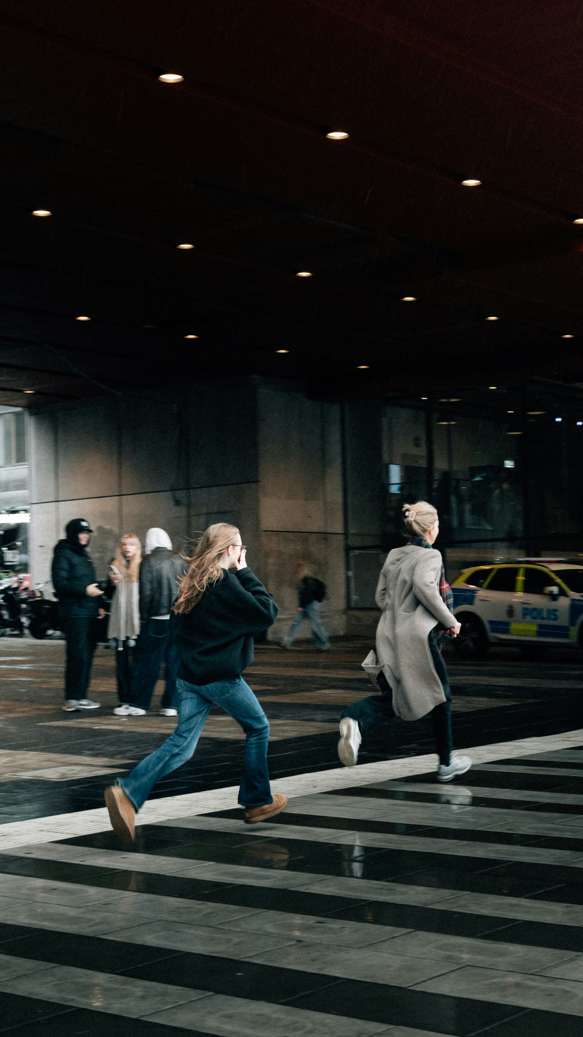 Duas mulheres atravessam uma faixa de pedestres com uma viatura de polícia próxima.