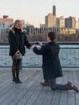 Man proposes to woman on pier with city skyline.