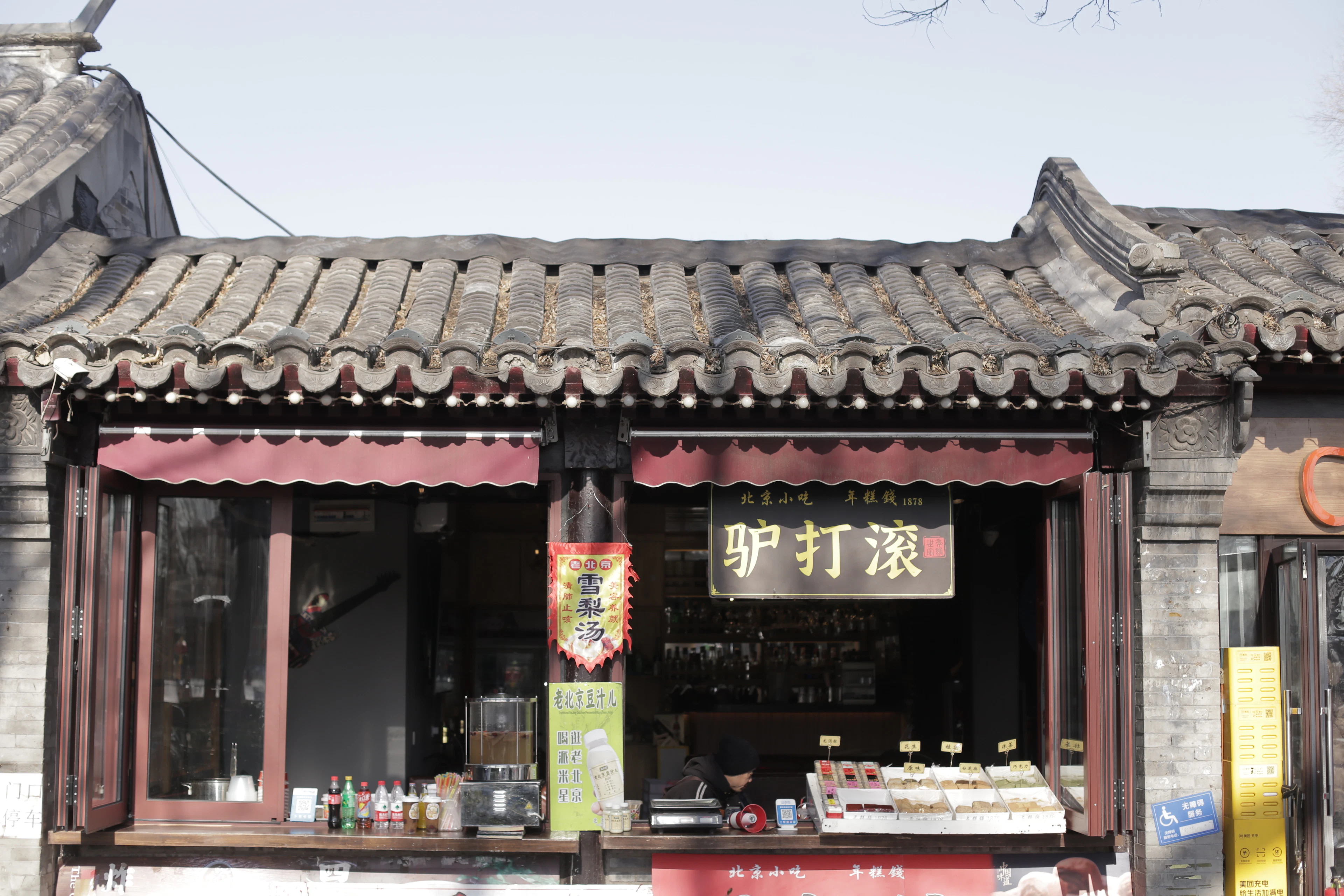 A traditional Beijing snack shop under a curved grey-tile roof, its lacquered sign advertising ludagun rolls above an open counter of trays of sweets and drinks.
