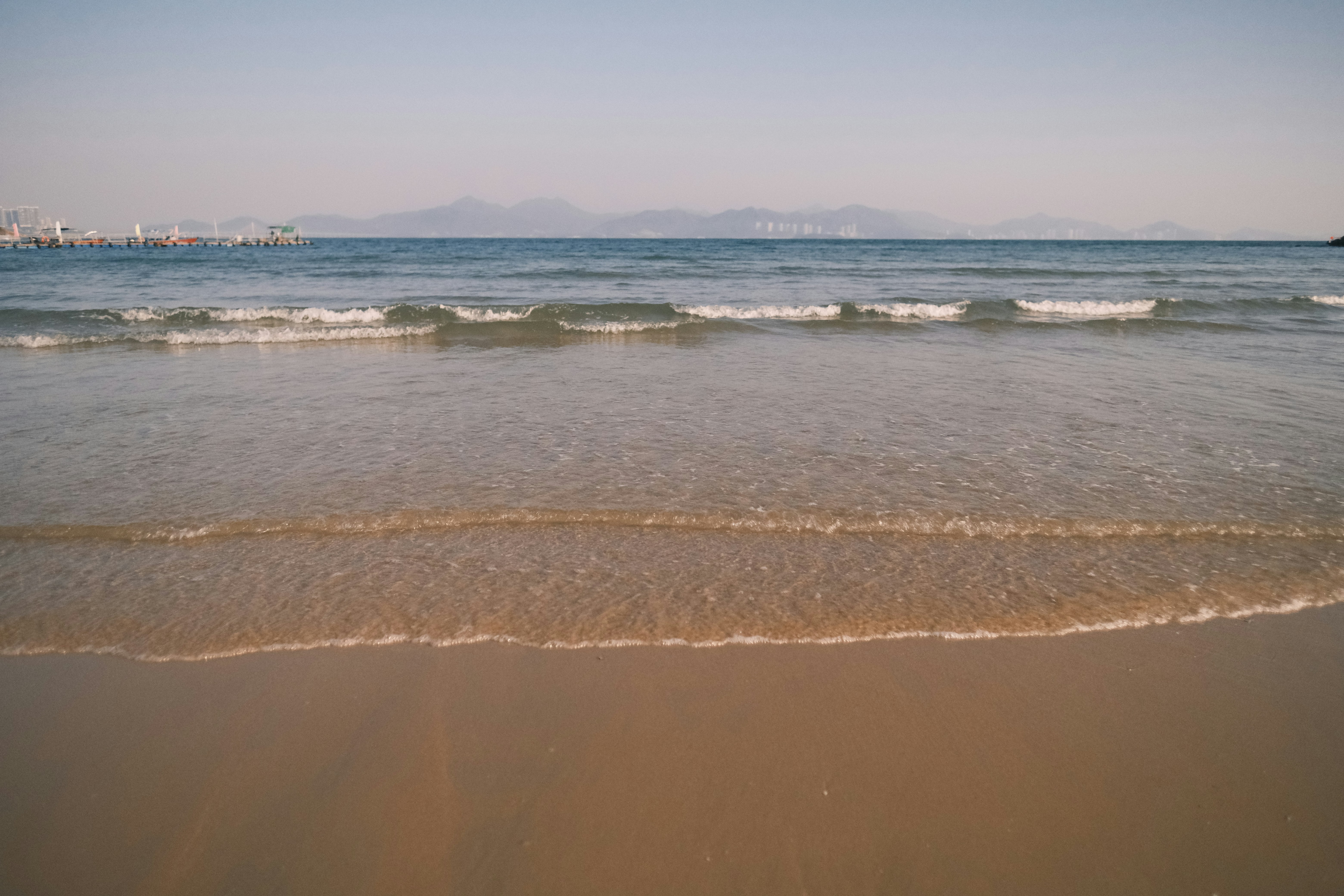 Olas suaves golpean una playa de arena con colinas lejanas.