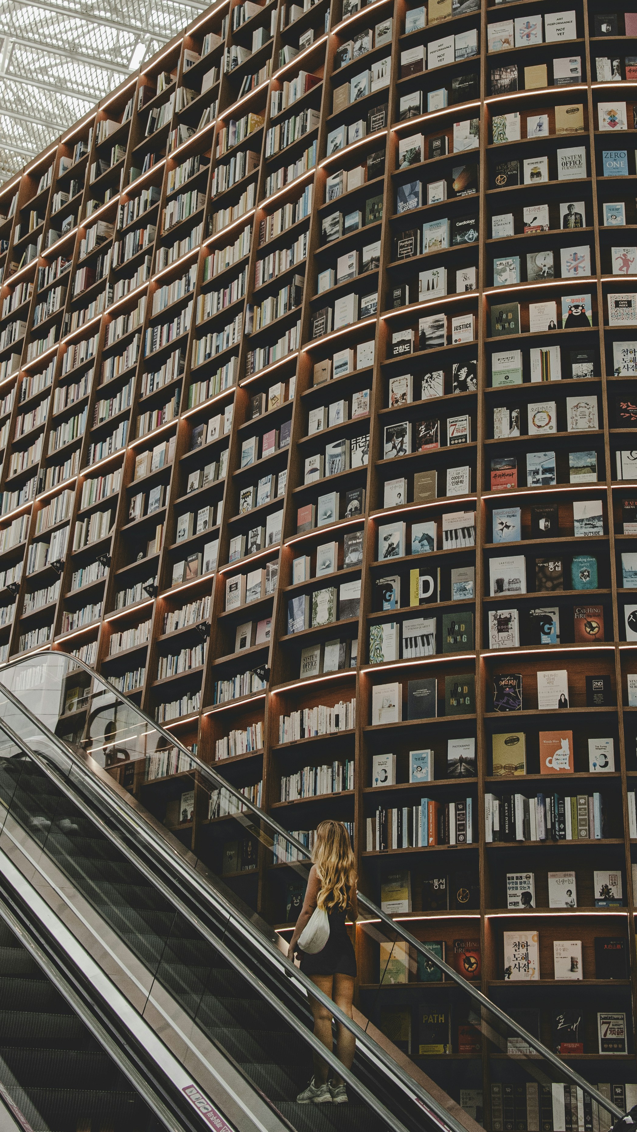 Une femme monte un escalator dans une vaste bibliothèque avec des étagères du sol au plafond.