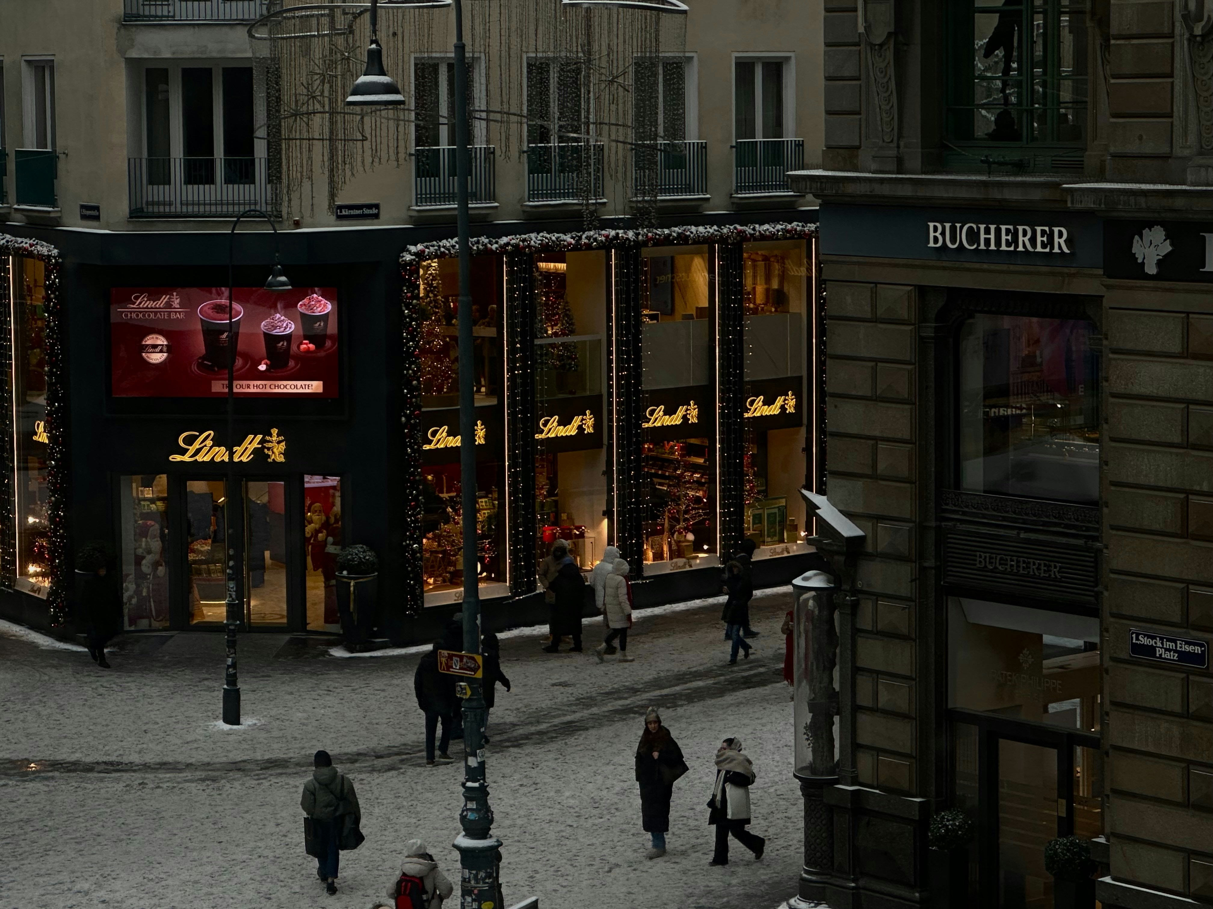 People walk on a snowy street past illuminated shops.