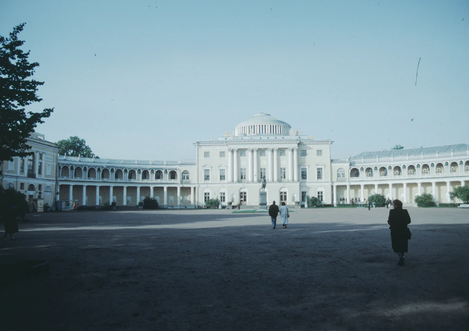The image depicts a large, white building with multiple levels and columns, reminiscent of classical architecture. The building is situated within a spacious courtyard, which appears to be empty at the moment. Two individuals can be seen walking towards the building in the foreground, adding a sense of scale to the scene.