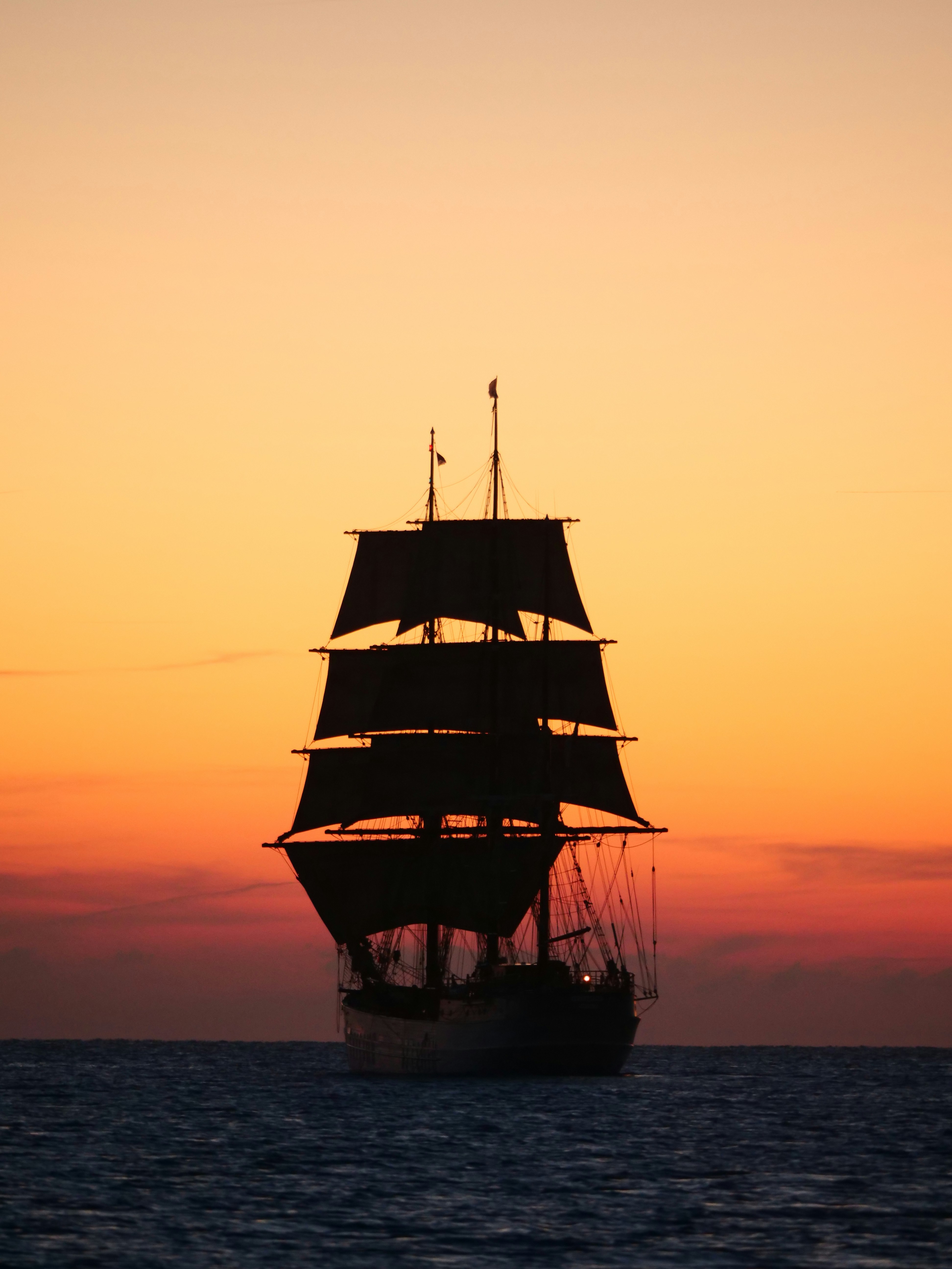 A tall ship sails on the ocean at sunset.