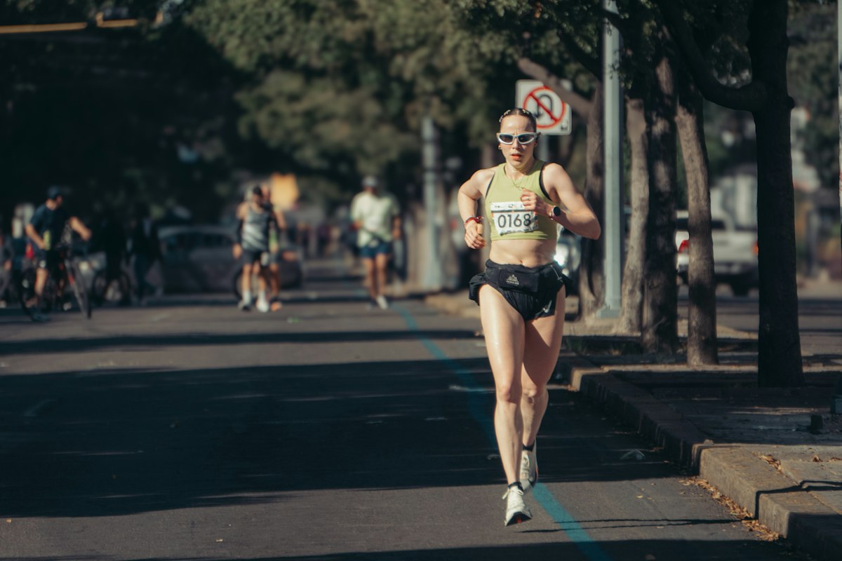 Coureuse avec dossard et lunettes de soleil en pleine course sur une avenue ensoleillée, d'autres coureurs en arrière-plan