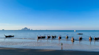 Longtail boats float on a calm ocean near a beach.