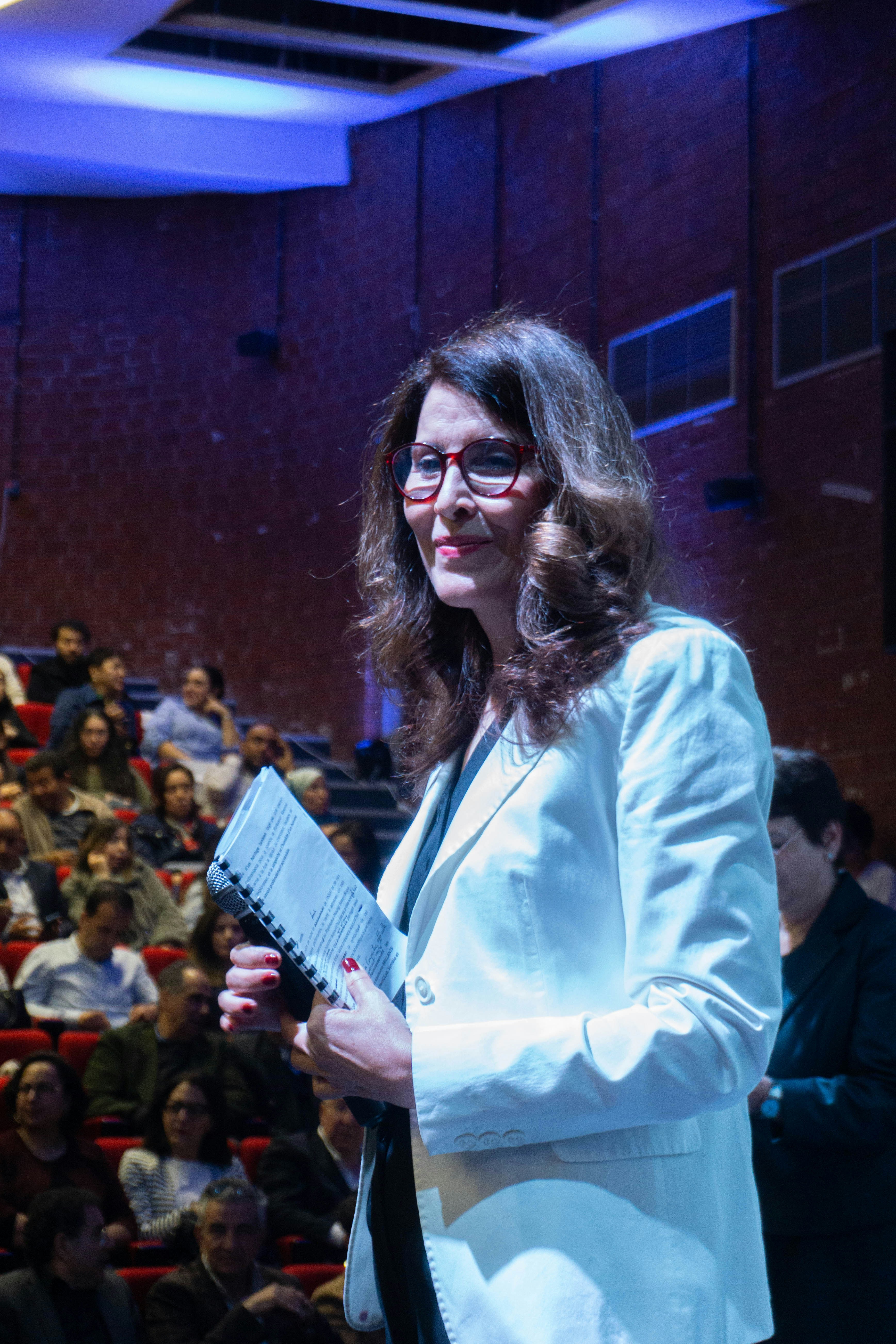 Mulher de blazer branco segurando um caderno no palco.